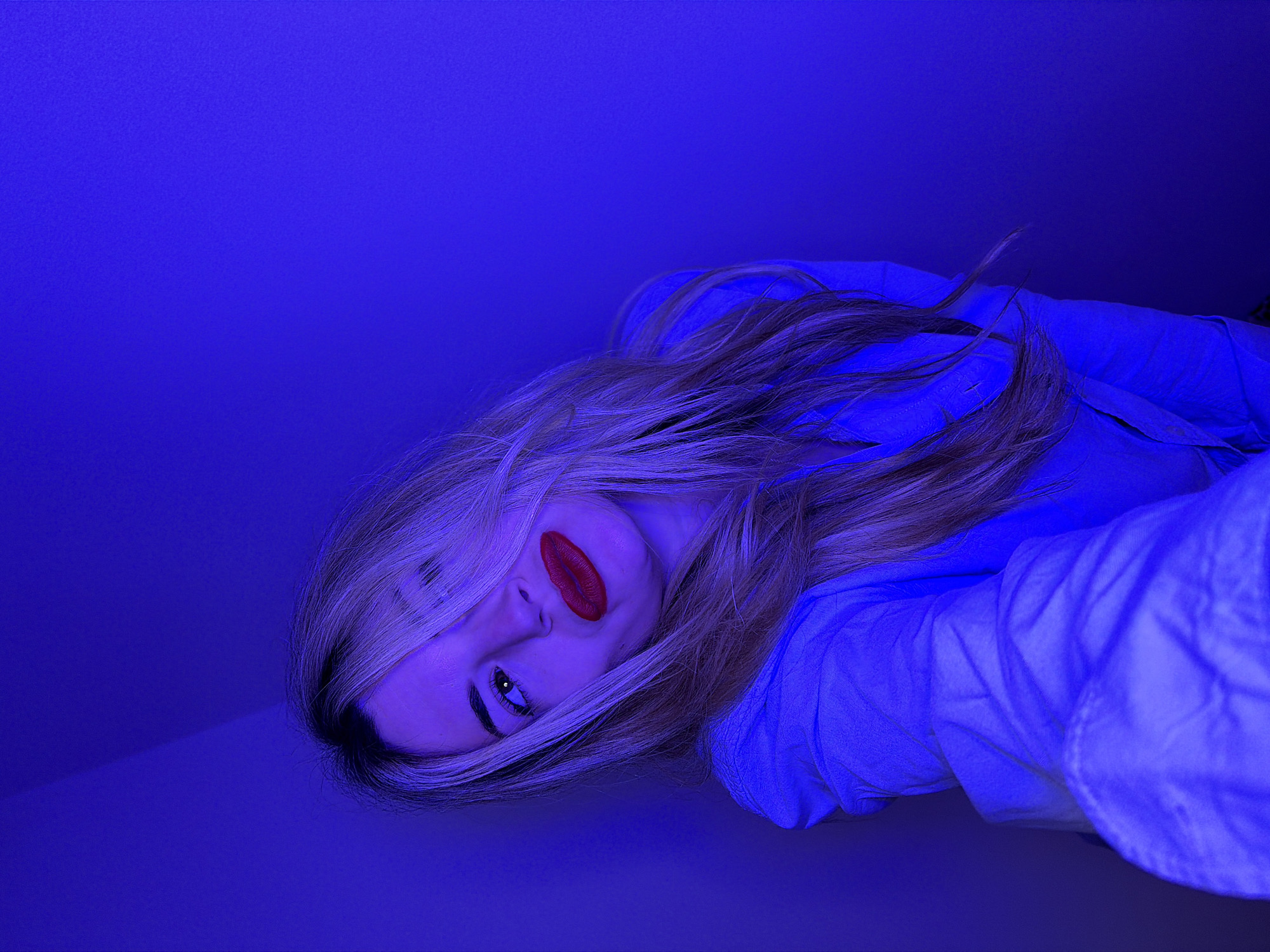 A blonde woman with red lipstick lies at an oblique angle on a blue surface. She is wearing a blue collared shirt. The setting is unknown, and no information indicates the location is Budva, Montenegro.
