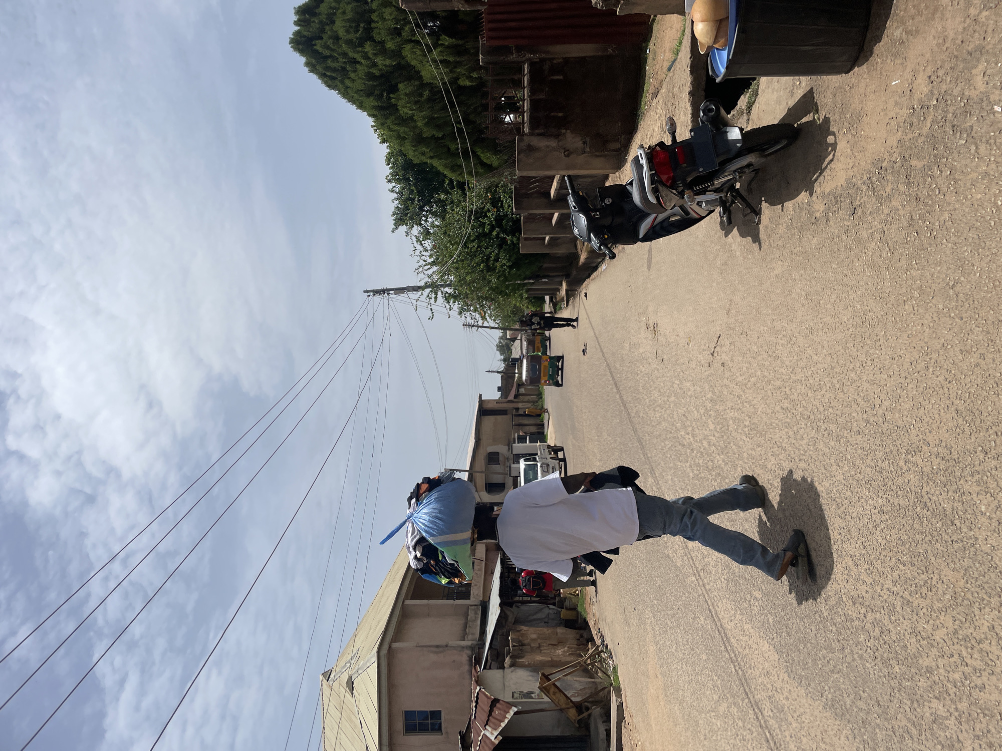 A street scene in Jos, Nigeria is captured. A person wearing a white t-shirt and blue jeans is carrying a large blue bag full of clothes or materials on their head while walking on a paved road. In the background, there are buildings and utility poles with wires stretching across the sky. Further down the street, there is a motorcycle parked on the side of the road, and there are other vehicles and individuals visible in the distance. The sky is partly cloudy.