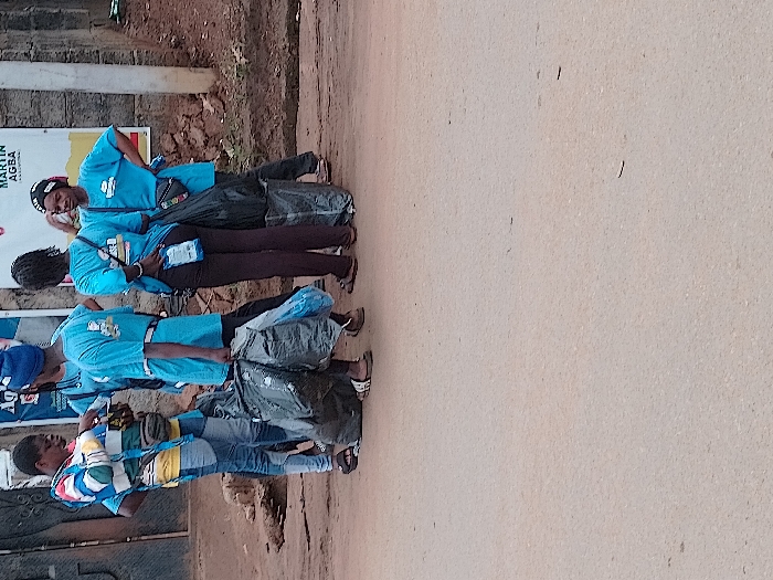 The scene in Orji, Nigeria, depicts six individuals, predominantly young adults, gathered on the unpaved shoulder of a light-colored, worn asphalt road. Five of the individuals wear light blue t-shirts with a white logo on the back, and some have dark trousers or jeans. Two individuals have white identification badges visible on their hips or waists. Several large black plastic trash bags, some appearing full, are present on the ground or being carried, suggesting a cleanup operation.
Specifically, one person on the far left wears a multi-colored long-sleeved shirt and blue jeans, facing right and interacting with another individual. A person with a blue beanie is partially visible, holding a black trash bag. One individual facing away carries a large black trash bag. Another person on the far right wears a black headwrap and stands with a hand on their hip, looking towards the right.
The background features a low, light-colored stone wall. Two large, faded banners are affixed to the wall, with visible text fragments including "AGBA" and "MARTIN". A dark metal gate is partially visible to the left of the group. The ground directly behind the individuals is uneven dirt. The scene is illuminated by natural daylight.