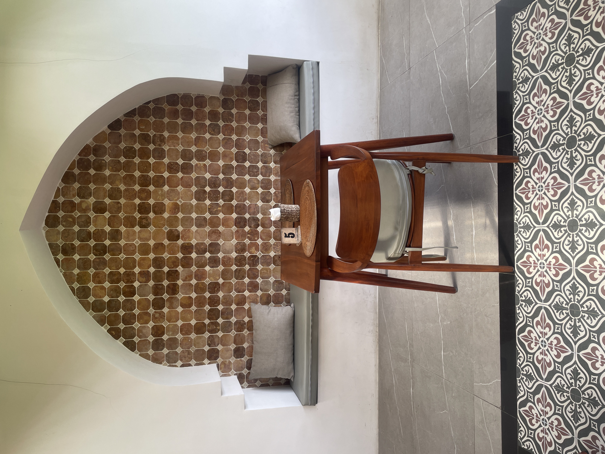 The image captures a cozy dining nook in Ubud District, Indonesia. The nook is defined by an arched recess in a white wall, the back of which is tiled with small, brown, octagon-shaped tiles arranged in a grid pattern. Two light grey pillows sit on a built-in ledge on either side of a small wooden table. On the table, there is a small decorative item and a number '5'. A single wooden chair with a white seat faces the table. The floor outside the nook is covered with large grey tiles with white veining, transitioning to decorative patterned tiles in black, white, and maroon near the edge of the frame. The overall impression is of a simple, elegant, and intimate dining space.