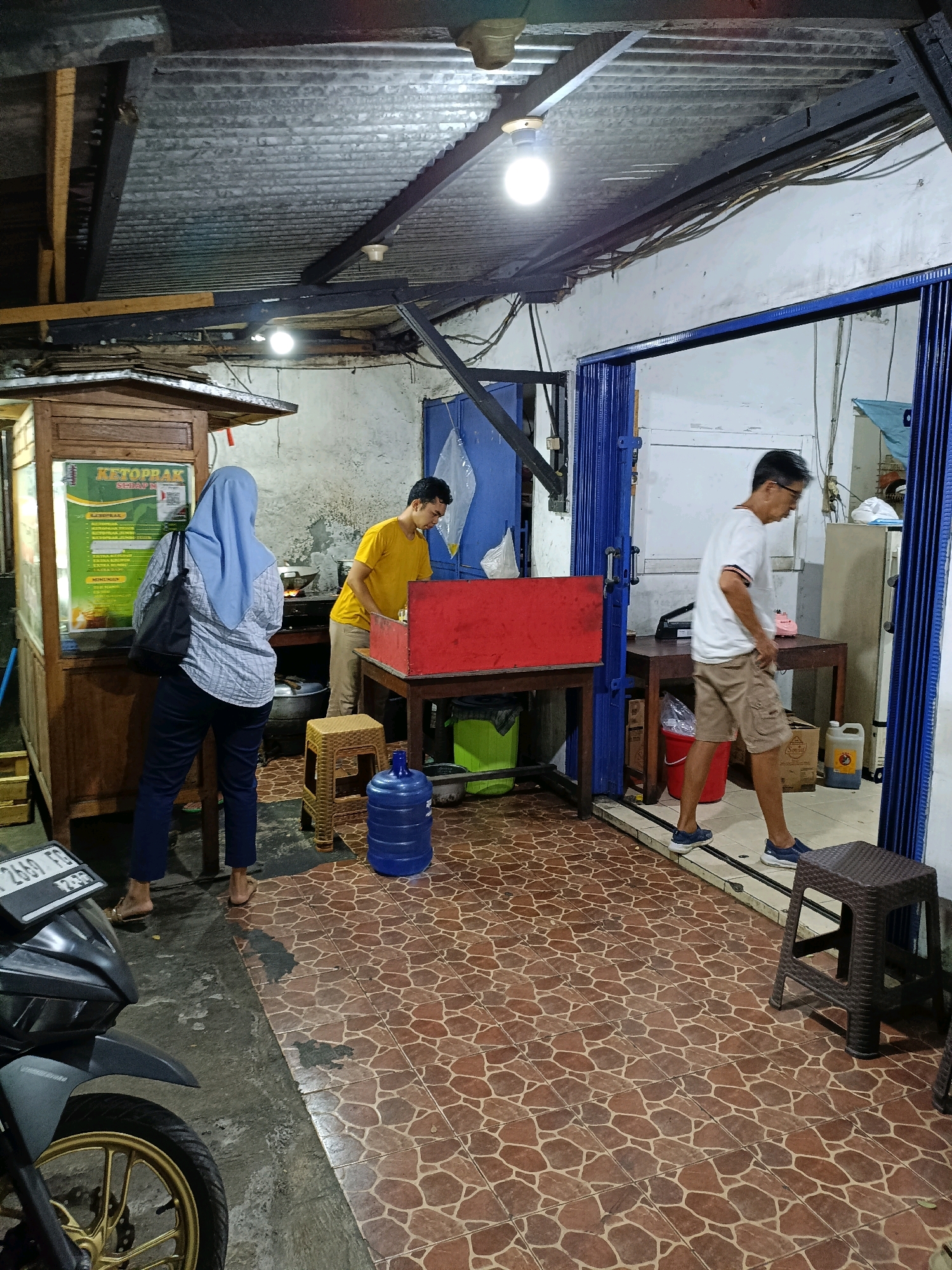In Maguwoharjo, Indonesia, a dimly lit outdoor food stall features a woman wearing a blue hijab and a checkered shirt, standing with her back to the camera, facing a menu on a wooden cart. To her right, a man in a yellow t-shirt is cooking. Another man in a white t-shirt and khaki shorts walks into a building with blue accordion doors, while a motorcycle is parked in the foreground. The area is paved with brown tiles and concrete, with a large blue water jug, a red food preparation station, and various tables and stools visible. The scene depicts a typical street food vending operation.