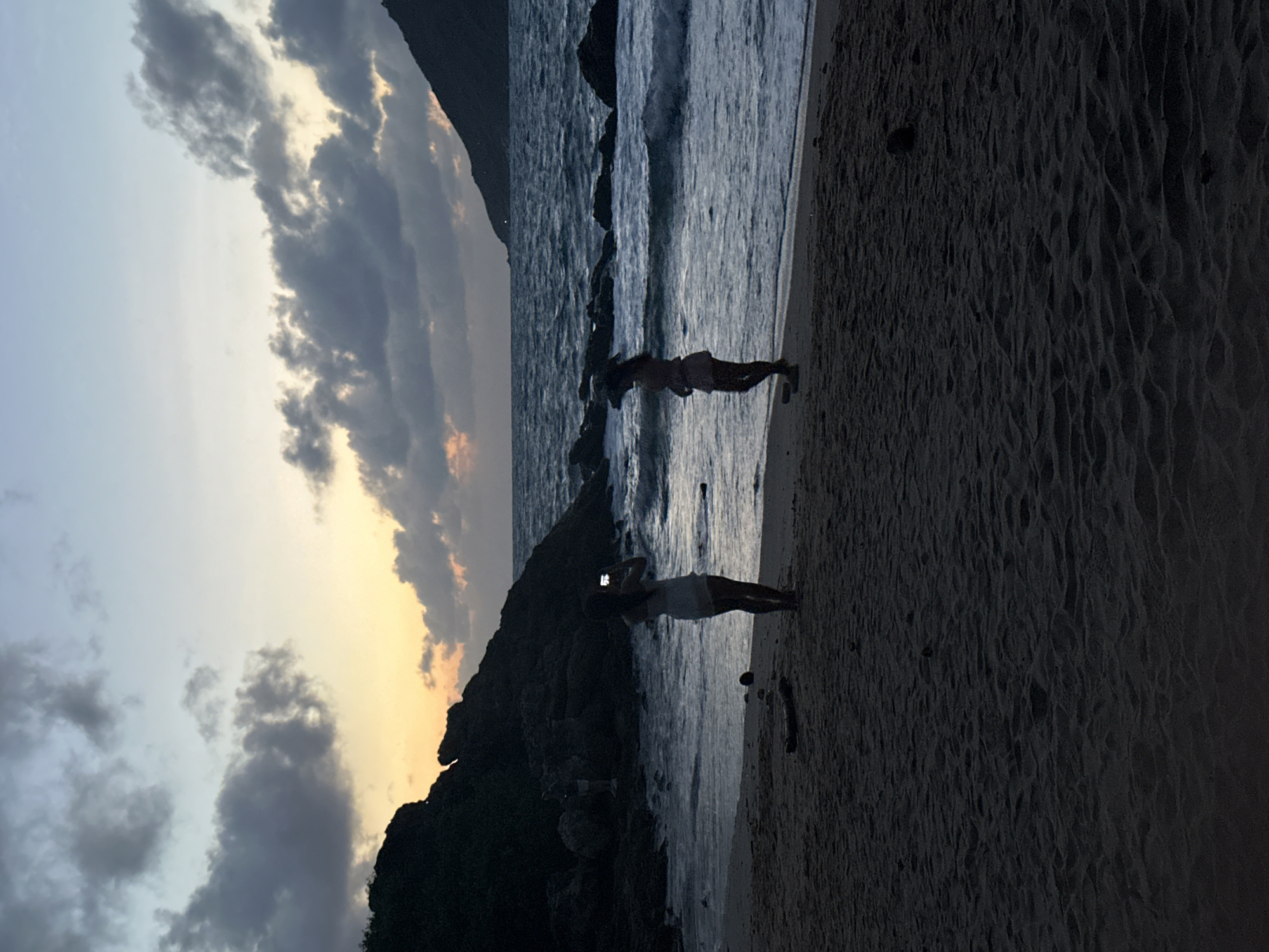 Two young women are silhouetted on a sandy beach in Rawai, Thailand, at dusk. The woman on the left is facing the ocean and holding a phone up to take a picture. She is wearing a light-colored dress. The woman on the right is also facing the ocean, with her back to the viewer. She is wearing a light-colored dress and appears to be looking at something in the distance. The ocean is choppy with waves breaking on the shore. Rocky outcrops are visible in the water and on the left side of the image, forming a cove. The sky is a gradient of pale blue, yellow, and gray with dark clouds, suggesting the sun has recently set. The beach is dark, with some debris visible in the sand.
