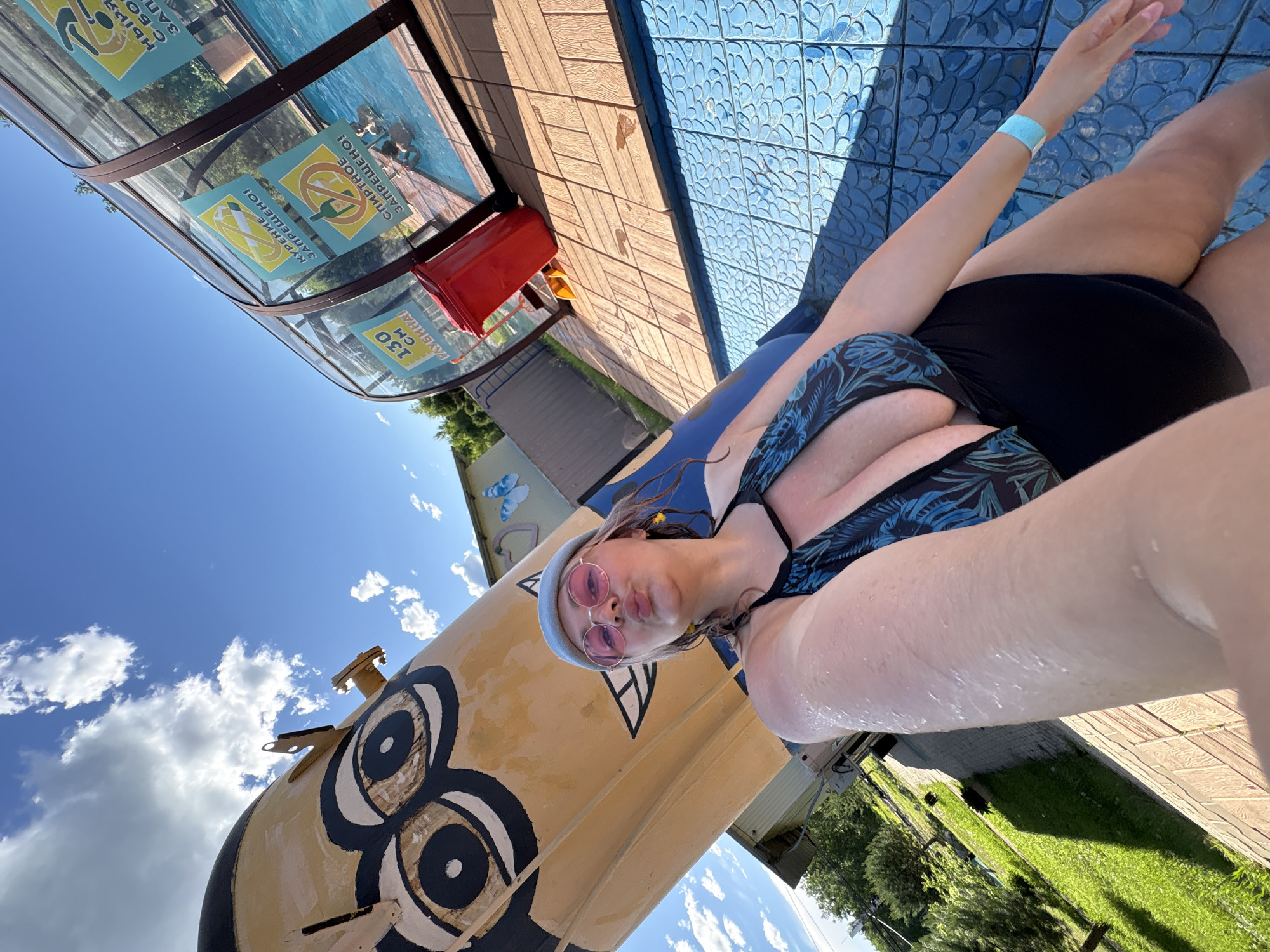 The image shows a woman in a black and blue floral patterned swimsuit and a light blue beanie, wearing pink sunglasses, posing for a selfie outdoors. She is in what appears to be an aquatic facility. To her left is a large, yellow cylindrical structure painted with the eyes of a cartoon character. Behind her, a glass-enclosed swimming pool area is visible, with several signs posted on the glass. One sign indicates a depth of 130 cm, and another shows a prohibition symbol over a bottle and glass. There are people swimming in the pool. A red trash can is also visible near the pool area. The ground around the pool is tiled with blue and brown wooden-patterned tiles. The background shows greenery, trees, and a bright blue sky with white clouds. The overall scene suggests a warm, sunny day at a water park or a similar recreational facility. The location is not specified in the image itself.