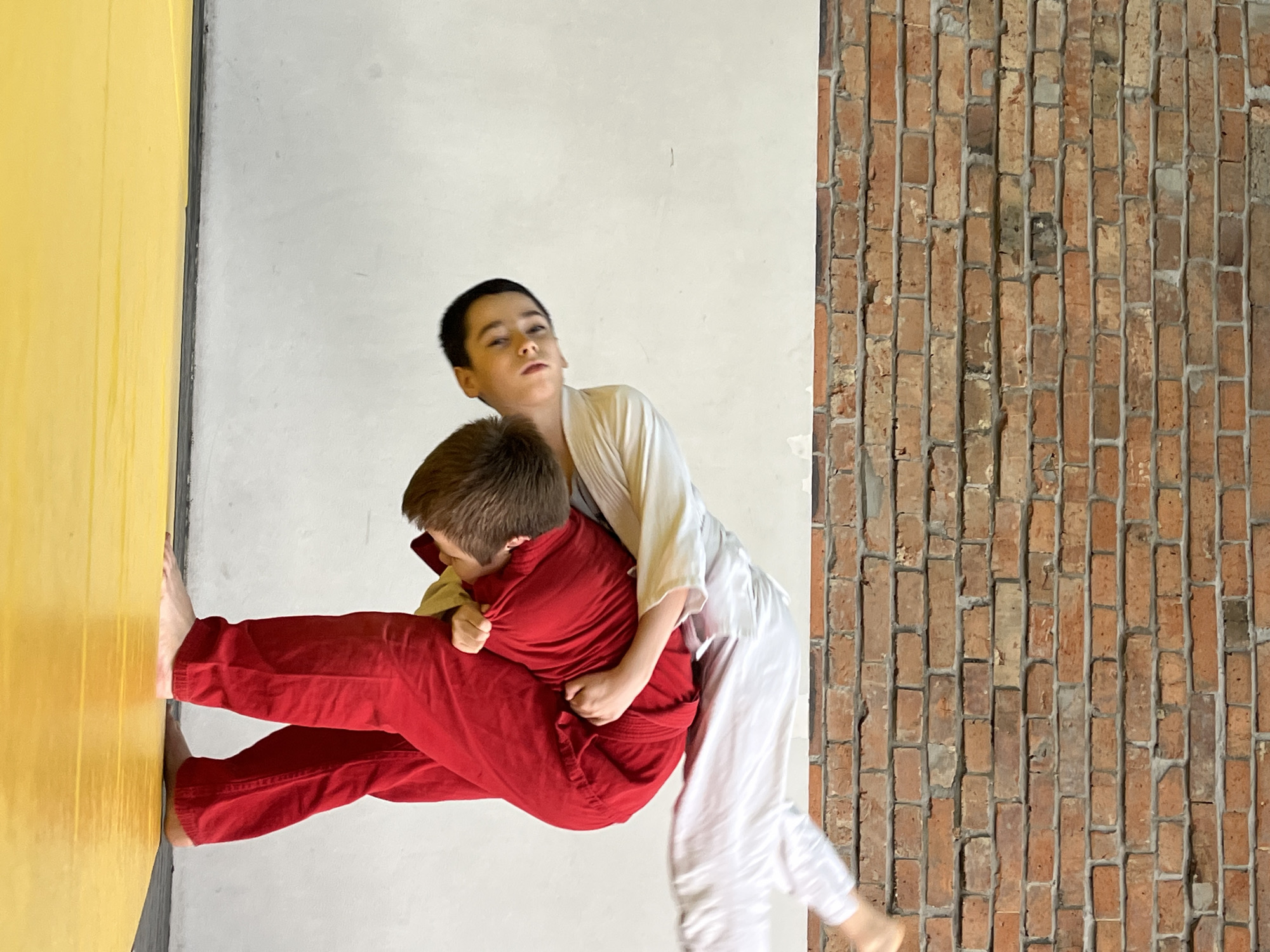 A full-shot image captures two young boys engaged in what appears to be a martial arts or grappling practice indoors. The scene is divided into three distinct background sections: a bright yellow surface on the far left, a plain white wall in the center, and an exposed brick wall on the right.
One boy, dressed in a white gi top and pants, stands on the right side of the frame. He has short, dark hair and looks towards the viewer with a focused, somewhat serious expression. He is actively holding the second boy in a grappling position.
The second boy is dressed in a red uniform, consisting of a red top and pants. He is positioned with his back mostly facing the viewer, revealing his light brown hair. His bare feet are planted against the bright yellow surface on the left, which could be a padded floor or a section of a wall being used for leverage. The boy in white has his arms wrapped around the red-clad boy's torso, seemingly performing a lift or a specific martial arts hold.
Both children are barefoot, indicating a training environment. The lighting is bright and even, suggesting an indoor setting with ample ambient light. The brick wall on the right features a rugged texture with variations in brick color, from orange to dark brown, and visible mortar lines. The overall scene depicts a dynamic moment of physical training within a dedicated studio or dojo. No visible text is present.