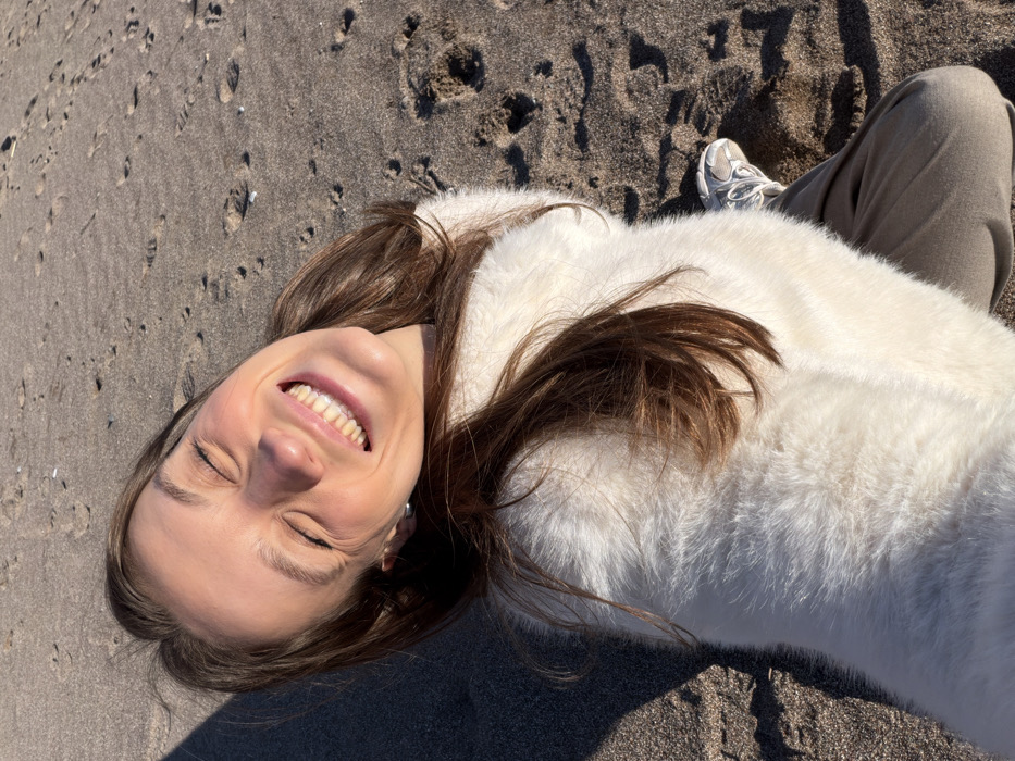 A woman lies on a sandy beach, angled towards the upper left. She wears a white, furry coat and tan pants, one leg bent slightly. Her brown hair spills onto the white fur. She smiles brightly with her eyes closed, facing the sunlight. The sand is dark and textured, with footprints visible. A white sneaker can be seen near her leg.