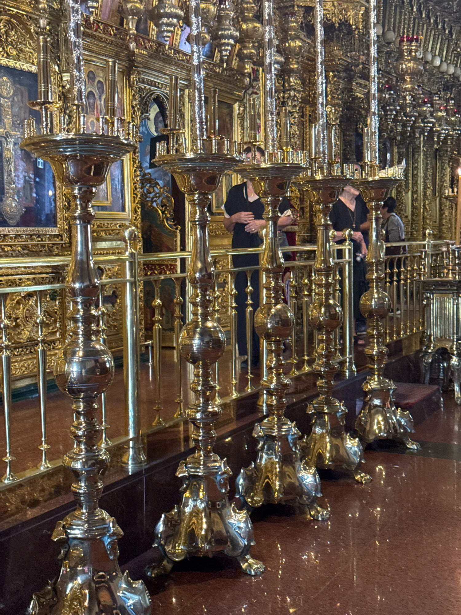 The image shows the interior of a richly decorated Orthodox church, likely in Tsakistra, Cyprus, given the historical context. The foreground is dominated by large, ornate golden candelabra, with intricate detailing and polished surfaces reflecting the ambient light. Behind the candelabra, a golden iconostasis is visible, adorned with religious icons. Several individuals, dressed in dark clothing, are standing in front of the iconostasis, seemingly observing the religious art or participating in a service. The floor appears to be polished marble or stone, further contributing to the opulent atmosphere. The lighting is warm and diffused, typical of interior church lighting, with some highlights glinting off the metallic surfaces. The overall impression is one of solemnity and grandeur.