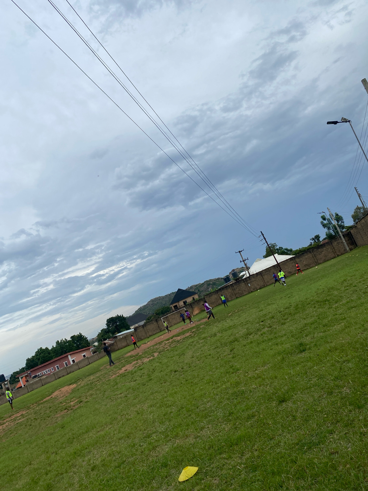 The image depicts a green soccer field with several individuals engaged in a game, located in Jos, Nigeria. The sky is overcast with dramatic clouds, and power lines stretch diagonally across the upper portion of the frame. A concrete wall borders the field, with buildings and trees visible beyond it. In the foreground, a yellow marker or cone lies on the grass. The players are scattered across the field, some actively participating in the game, while others appear to be observing. The overall atmosphere suggests a casual sporting event taking place in an urban environment.