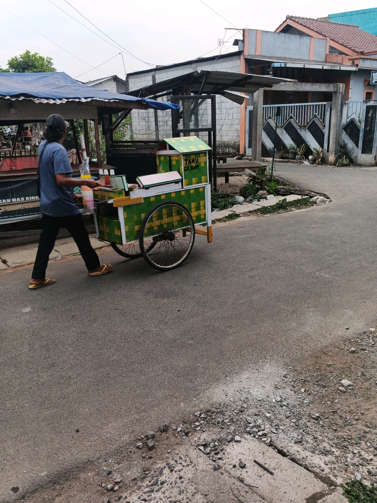 The image shows a man pushing a cart on a paved road in Kadu, Indonesia. The man is wearing a blue t-shirt and dark pants, with yellow flip-flops. He is walking from left to right, facing away from the camera. The cart is green and yellow, with two large wheels. It appears to be a food cart, with containers and bottles visible on top. In the background, there are houses and buildings, some with corrugated metal roofs. There is also a blue tarp covering part of a structure. The road is asphalt and shows some signs of wear and tear. The sky is overcast.