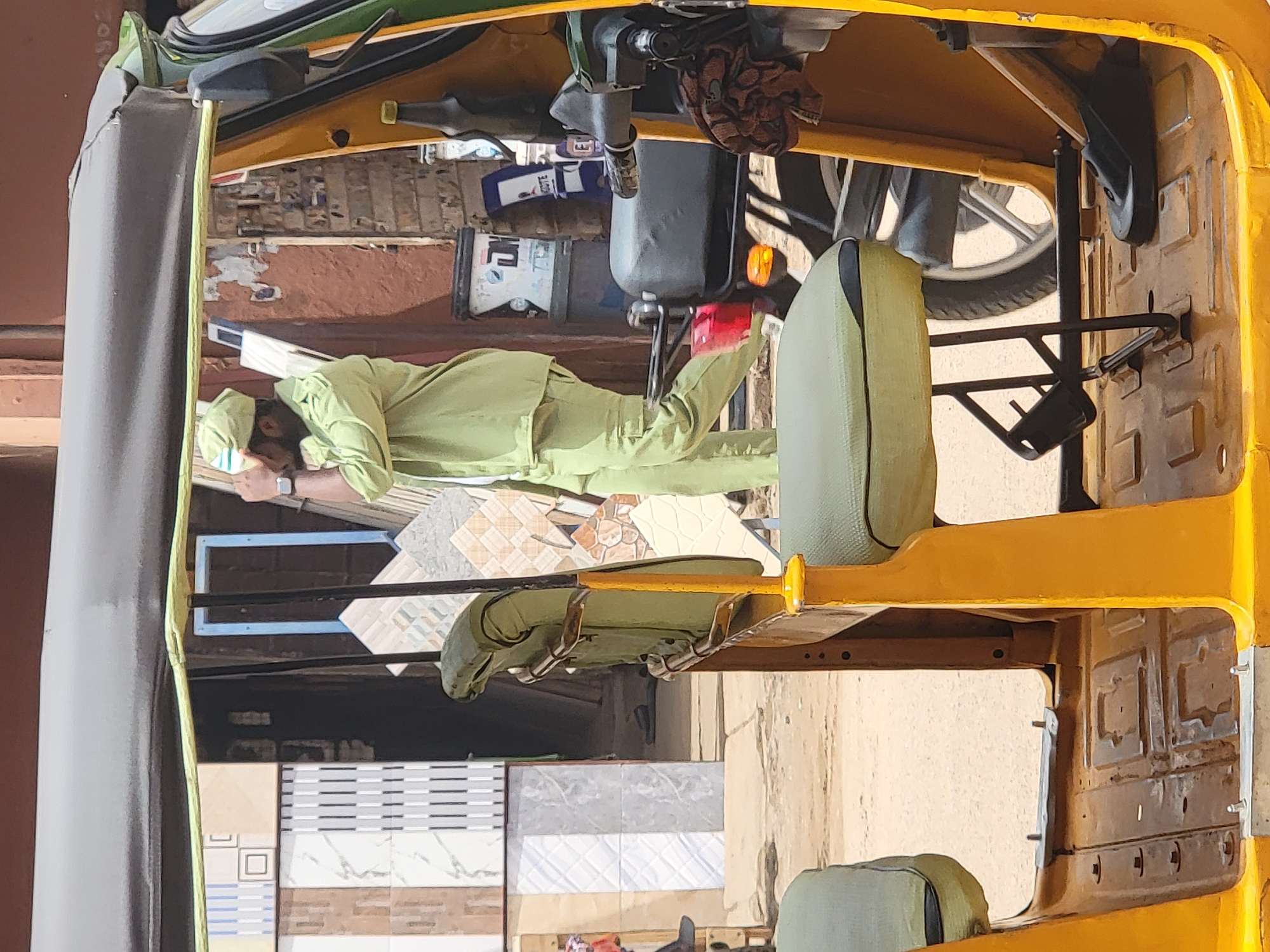 A man wearing a light green uniform and a cap is lying down inside the open back of a yellow three-wheeled vehicle, likely a type of auto rickshaw common in some regions. The vehicle is filled with tiles, with stacks of them visible in the background. The location is presumed to be Maiburiji, Nigeria.
