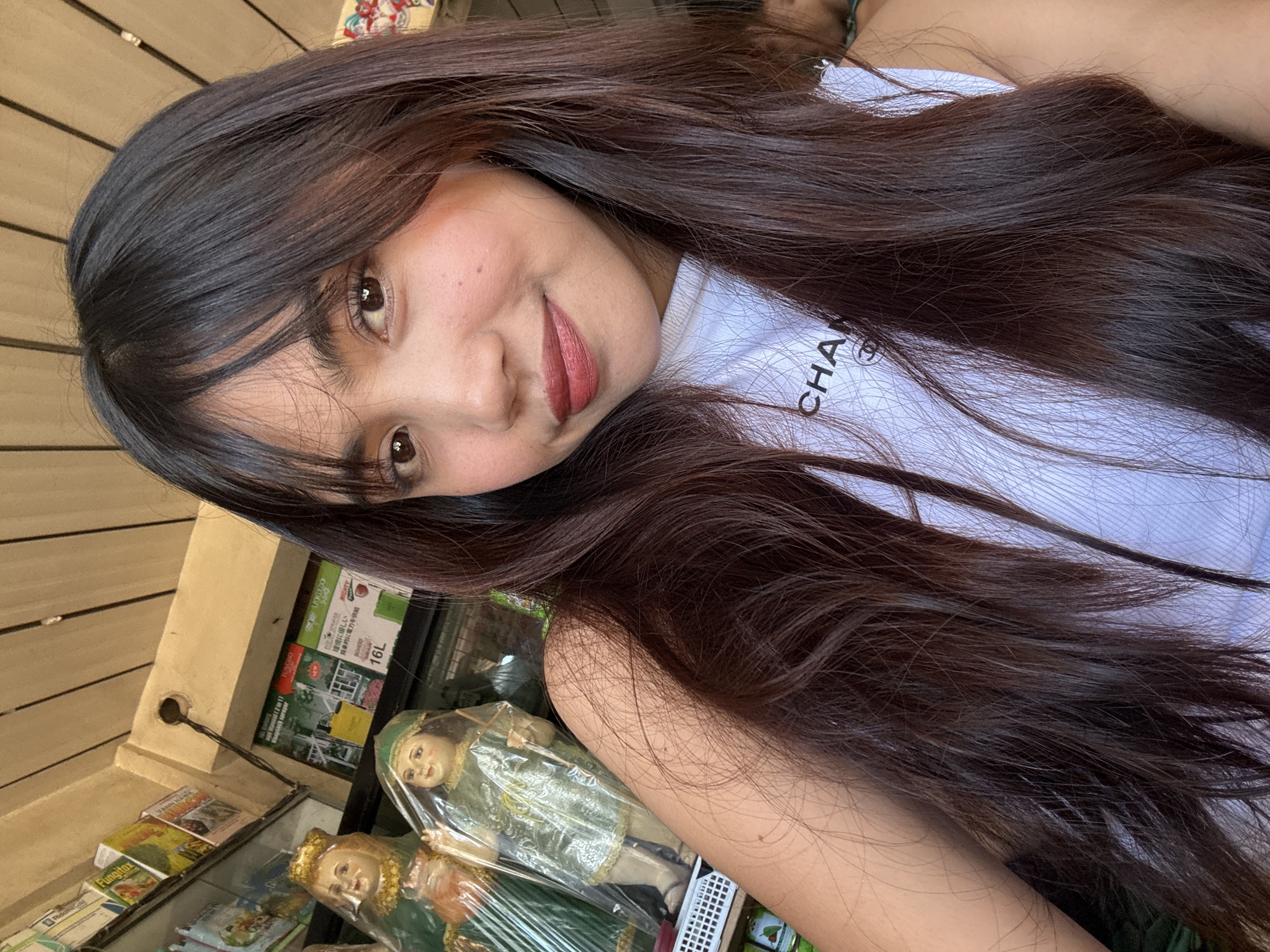 A young woman with long, dark hair is taking a selfie in what appears to be a small shop in Tagum City, Philippines. She is wearing a white tank top with a black logo that reads "CHANEL". In the background, several statues are visible, wrapped in plastic. One statue on the left is larger and wears a gold crown and red garment. Another statue to its right wears a green cap and gown. Shelves with various items, possibly agricultural products or general goods, are visible behind the statues. The ceiling above is constructed of wooden planks. The lighting suggests an indoor environment with natural light filtering in from above.