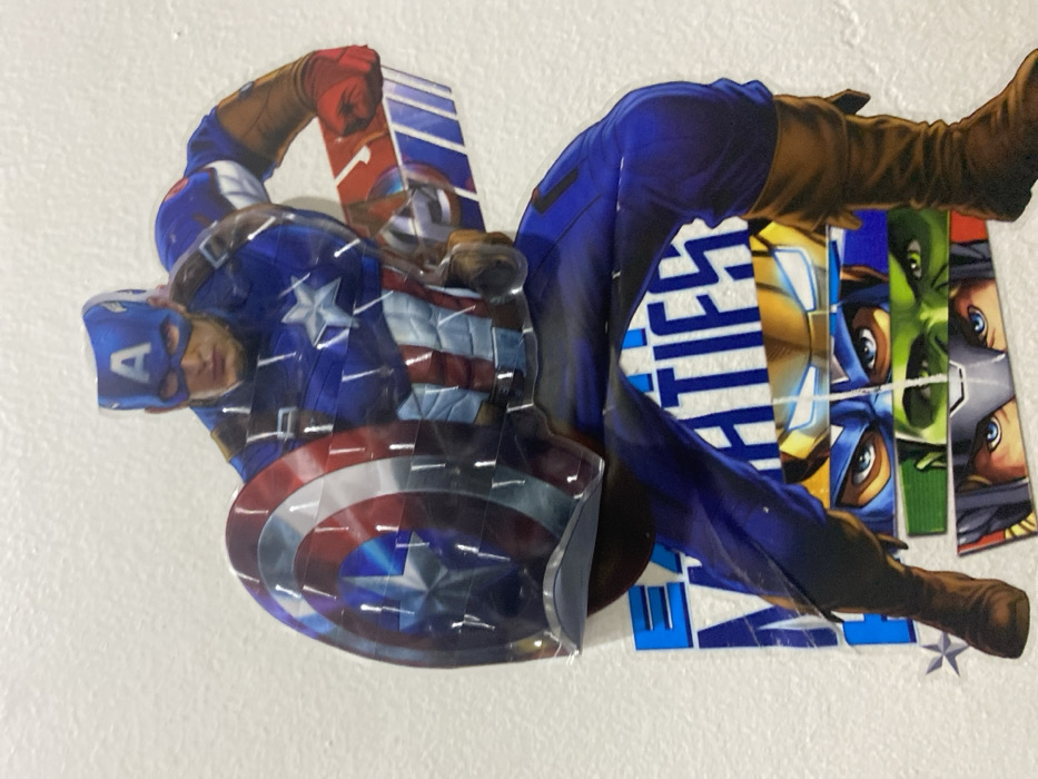 This image captures a multi-layered graphical display affixed to a textured, off-white wall in Tema, Ghana. The primary subject is a large, cut-out depiction of Captain America in a dynamic, crouching stance, facing forward and slightly to the viewer's left. He wears his iconic blue uniform with a white star on the chest, red and white stripes on the abdomen, and brown boots and gloves. His blue mask features a white "A" on the forehead, and he holds his circular red, white, and blue vibranium shield in his left hand. The graphic material appears to be a glossy print, exhibiting reflections from an ambient light source, particularly on the shield and other uniform elements.
Behind Captain America's lower body, partially obscured, are stacked horizontal strips featuring close-ups of the masked faces or eyes of other Marvel characters. From top to bottom, these appear to be Iron Man (gold and red mask with glowing blue eyes), Captain America (blue mask with exposed eyes), Hulk (green face with intense eyes), and Thor (grey helmet with exposed eyes and blonde hair). Below Captain America's midsection, the text "MATIFES" (or similar incomplete word) is visible in blue and white lettering, positioned on a white background, suggesting a larger banner or sign underneath the characters. The entire graphic unit is a singular, assembled display mounted directly onto the wall. The wall surface itself has a subtle, uneven texture, likely painted plaster or concrete.