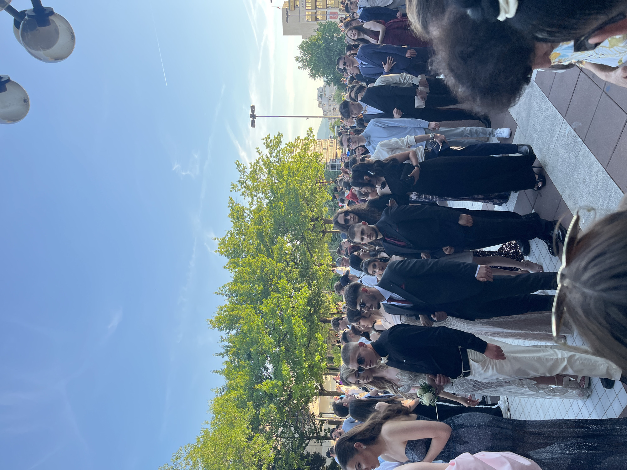 A large group of people dressed in formal attire stands outdoors in Blagoevgrad, Bulgaria. The crowd, presumably attending a graduation or formal event, is lined up. Most individuals are in suits and dresses. A large green tree stands to the left of the group, and buildings are visible in the background. The sky is clear and blue. A light pole with lamps is also visible.