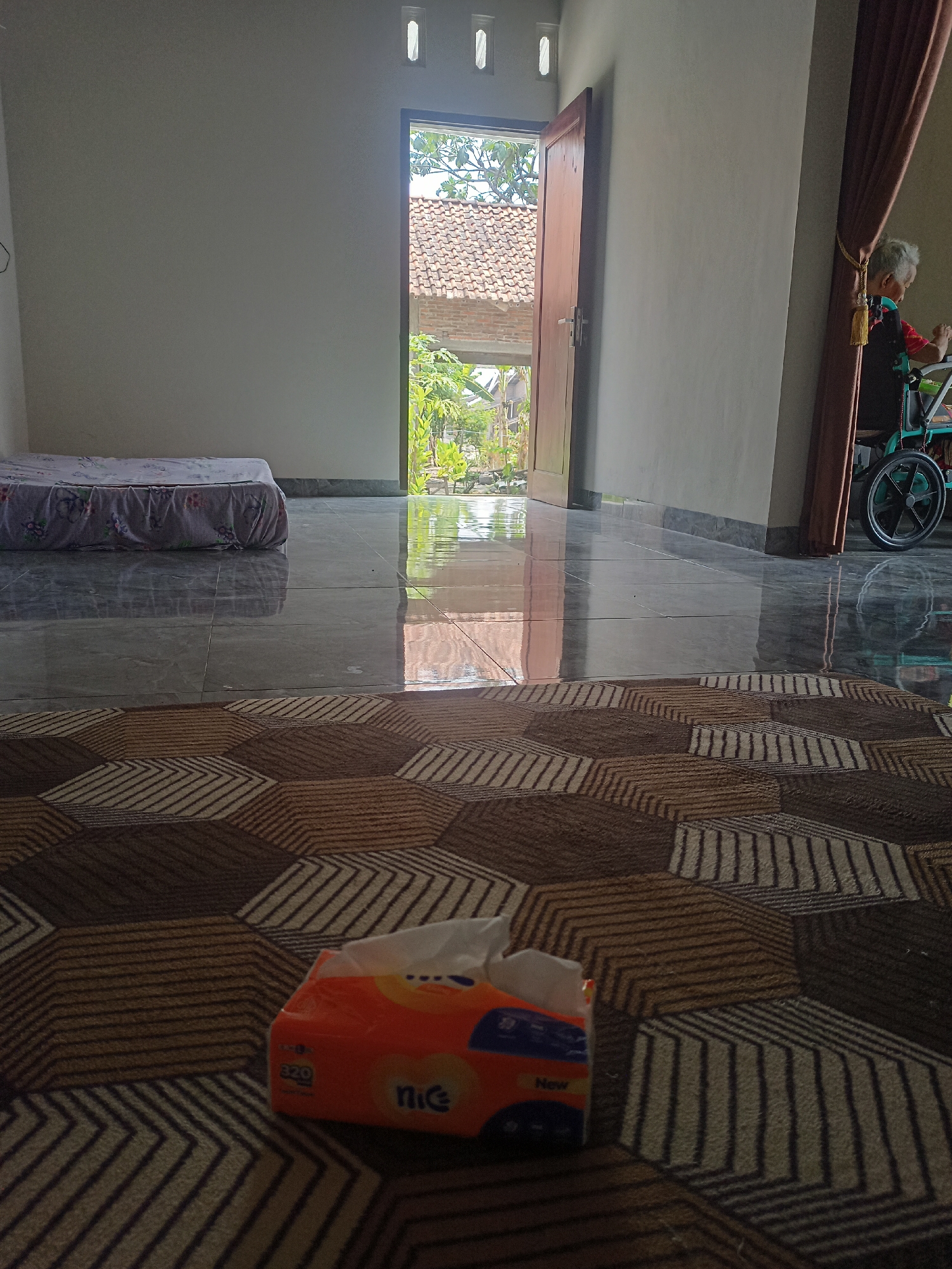 The image depicts an indoor scene, likely a room in a house, with an open doorway leading to an outdoor area. The room has a tiled floor with a reflective surface, and a patterned carpet in the foreground. A mattress is visible on the left side of the room, and a box of tissues sits on the carpet. In the background, through the doorway, there is a view of a tiled roof and lush green vegetation, characteristic of a rural or suburban environment like Karangtalun, Indonesia. A person is partially visible on the right side of the frame, seated in a wheelchair near a curtained window. The lighting suggests it is daytime.