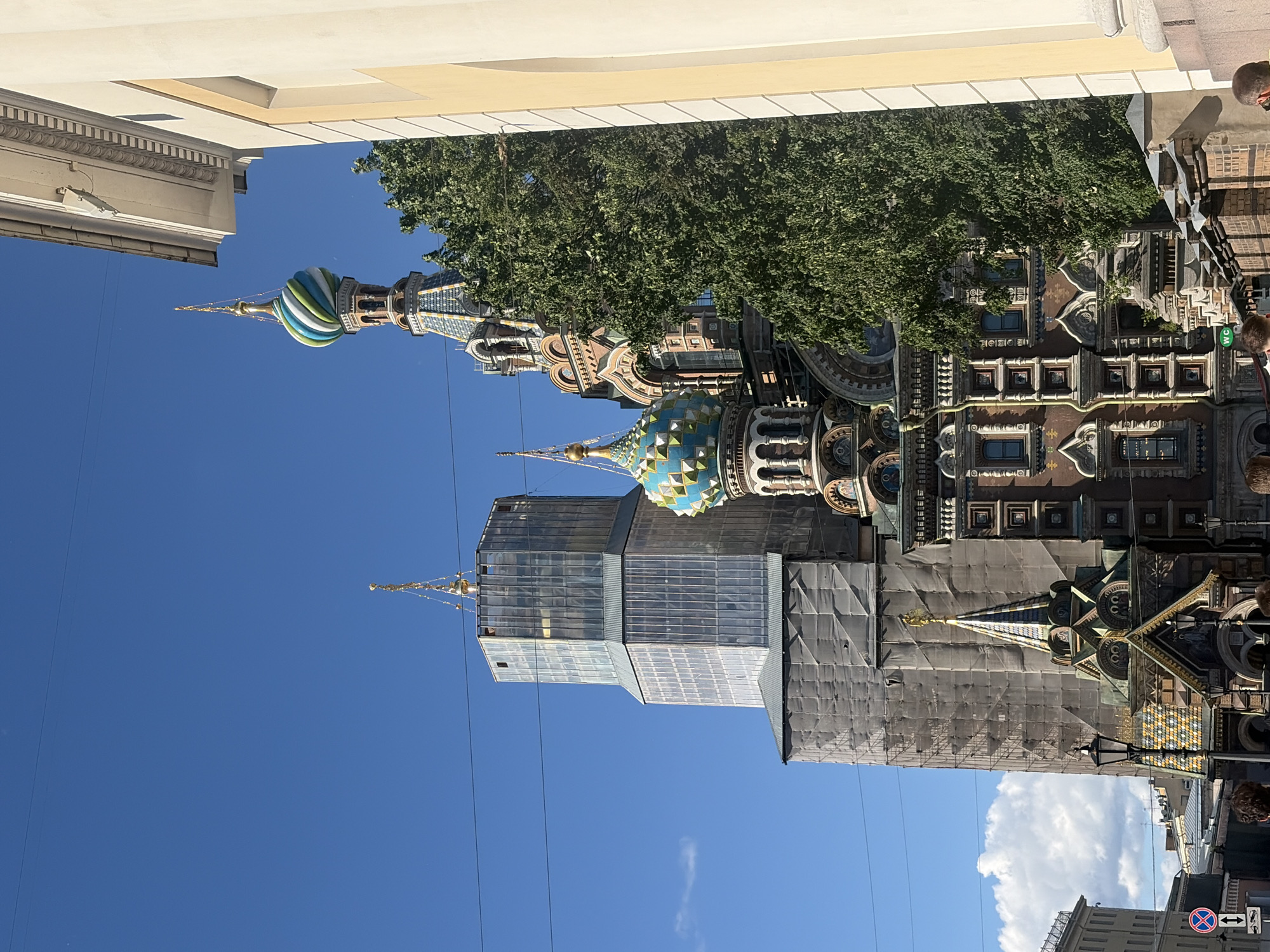 The image depicts a street scene in Saint Petersburg, Russia, with a prominent view of the Church of the Savior on Spilled Blood. The architecture is ornate, featuring colorful domes and intricate details. The church is partially obscured by scaffolding and a modern glass building that appears to be under construction or renovation. Lush green trees frame the upper portion of the church. The sky is a clear, vibrant blue, suggesting a sunny day, likely in the late morning or early afternoon given the bright light. The low angle of the sun casts some shadows, but the overall atmosphere feels bright and pleasant.
Several people are visible in the lower right of the frame, walking along the street, adding a sense of everyday activity to the historic setting. One individual is wearing sunglasses and appears to be looking up at the church. Another person is partially visible, with their back to the camera. There are street signs visible, including a traffic sign with a red circle and a horizontal bar, indicating "no entry" or a similar restriction, and a sign with a WC symbol, denoting a restroom. The presence of overhead wires suggests an urban environment with active infrastructure. The overall scene captures a blend of historical grandeur and ongoing development in a European city.