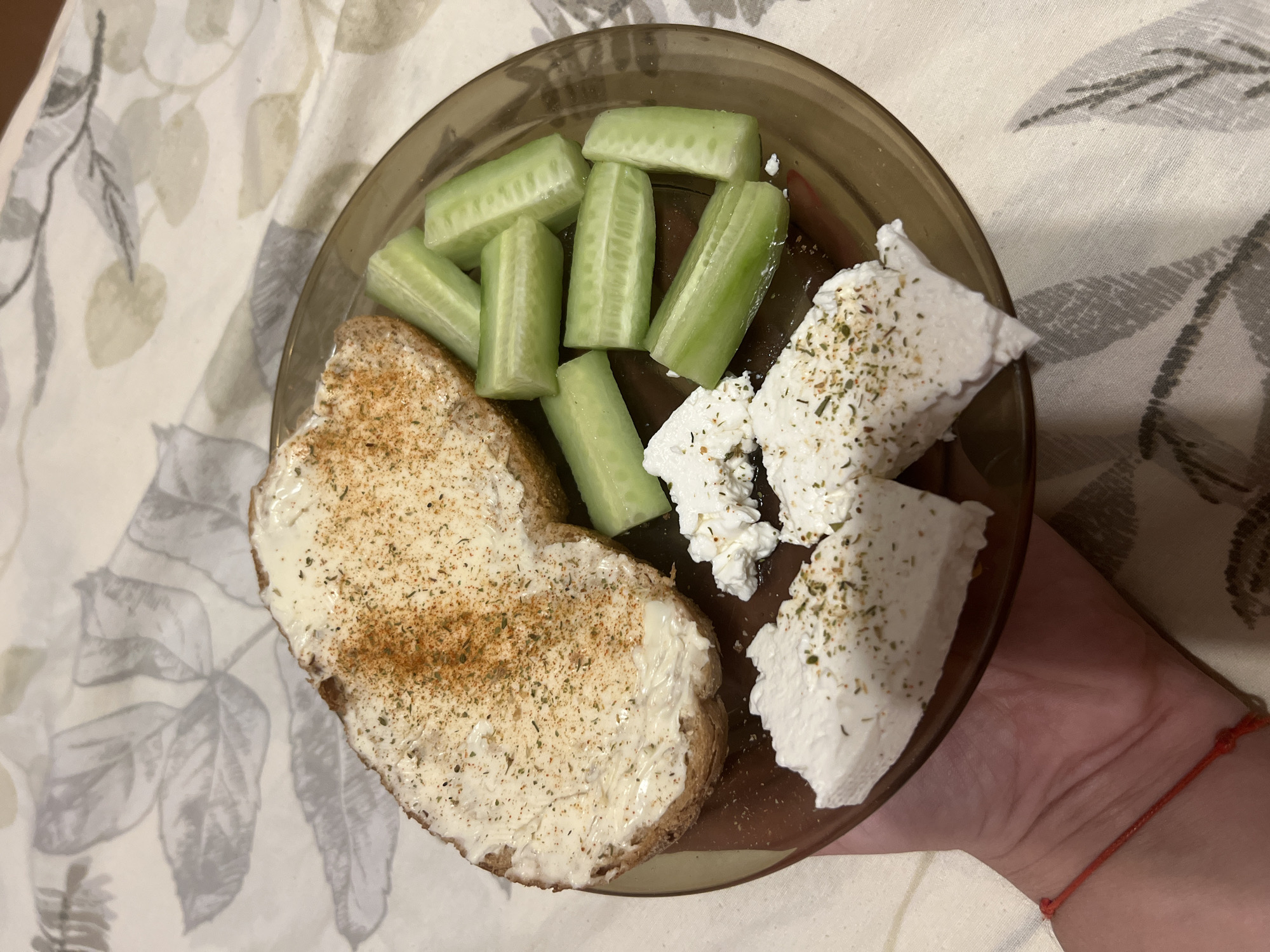 A plate containing a slice of bread with butter and spices, cucumber slices, and crumbled cheese is held in a person's hand. The plate is brown and sits on a tablecloth with a leaf pattern. The background is blurred. The food appears to be part of a meal, likely consumed indoors. The image was taken in Vratsa, Bulgaria.