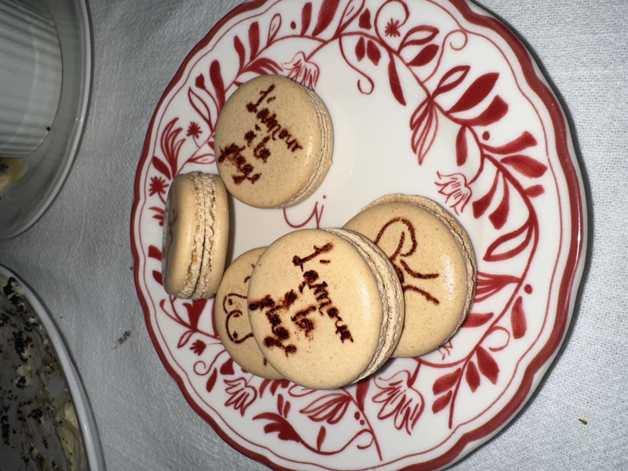 A close-up, overhead shot displays several macarons arranged on a decorative plate, set against a light-colored, possibly textured tablecloth. The primary subjects are five light-brown macarons, some of which bear dark brown, hand-drawn text or designs on their top shells.
One macaron prominently features the French phrase "L'amour à la page" (Love on the page or fashionable love), written in a stylized script. Another macaron also clearly shows "L'amour," while others have partial or less discernible markings. The macarons exhibit their characteristic "feet" at the base.
They are resting on a round, white ceramic plate adorned with a vibrant red, intricate border pattern consisting of floral and leafy motifs. To the left, part of a clear glass dish or container is visible, containing a light-colored, creamy dessert with dark speckles, possibly chocolate or coffee, suggesting another sweet treat.
The scene conveys an intimate dessert setting, likely indoors under artificial lighting. The overall impression is one of artful presentation, possibly for a special occasion, within a home or restaurant in North Kuta, Indonesia. No people are visible, and the activity implied is the enjoyment or serving of these confections.
