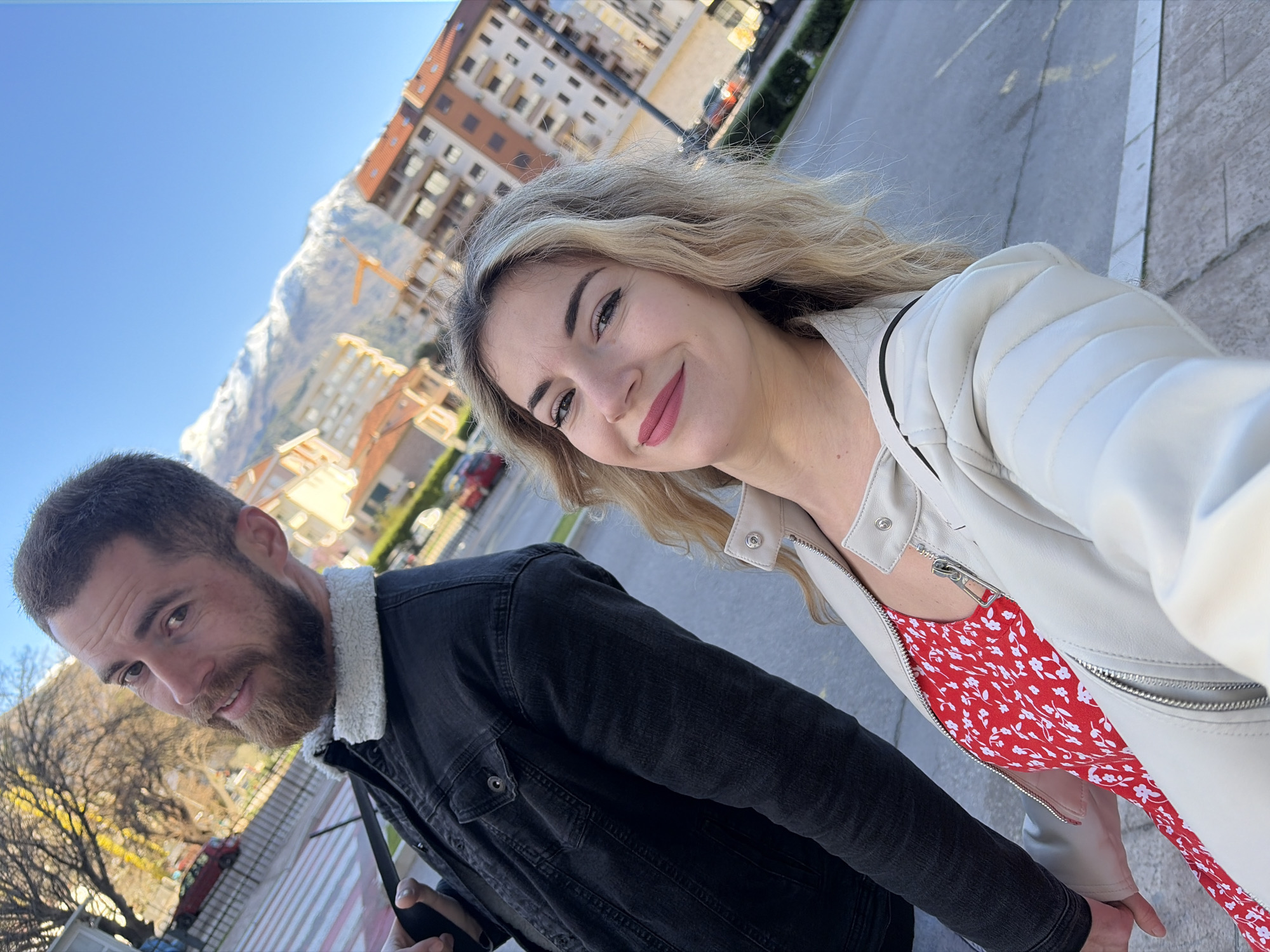 A selfie-style portrait taken outdoors in Budva, Montenegro, features two individuals: a woman in the foreground and a man positioned behind and to her left. The woman, with blonde wavy hair and light skin, wears a cream-colored leather-look jacket over a red dress patterned with small white flowers. She smiles towards the camera, displaying pink lipstick and dark eyeliner. Her right hand is visible in the lower left portion of the frame, holding the man's left hand. The man has short brown hair and a brown beard, and is wearing a dark denim jacket with a white sherpa-style collar. He also smiles slightly towards the camera. His right hand is partially visible near his waist, holding a dark object. The background shows an urban street scene with a paved road and a sidewalk. Multi-story buildings with various architectural styles, including some with reddish-orange tiled roofs, line the street. In the far background, prominent snow-capped mountains are visible under a clear, bright blue sky. A yellow construction crane is present among the buildings, situated in front of the mountains. Several red vehicles are partially visible in the mid-ground. Deciduous trees with bare branches and a dark metal fence are on the left side of the frame.
