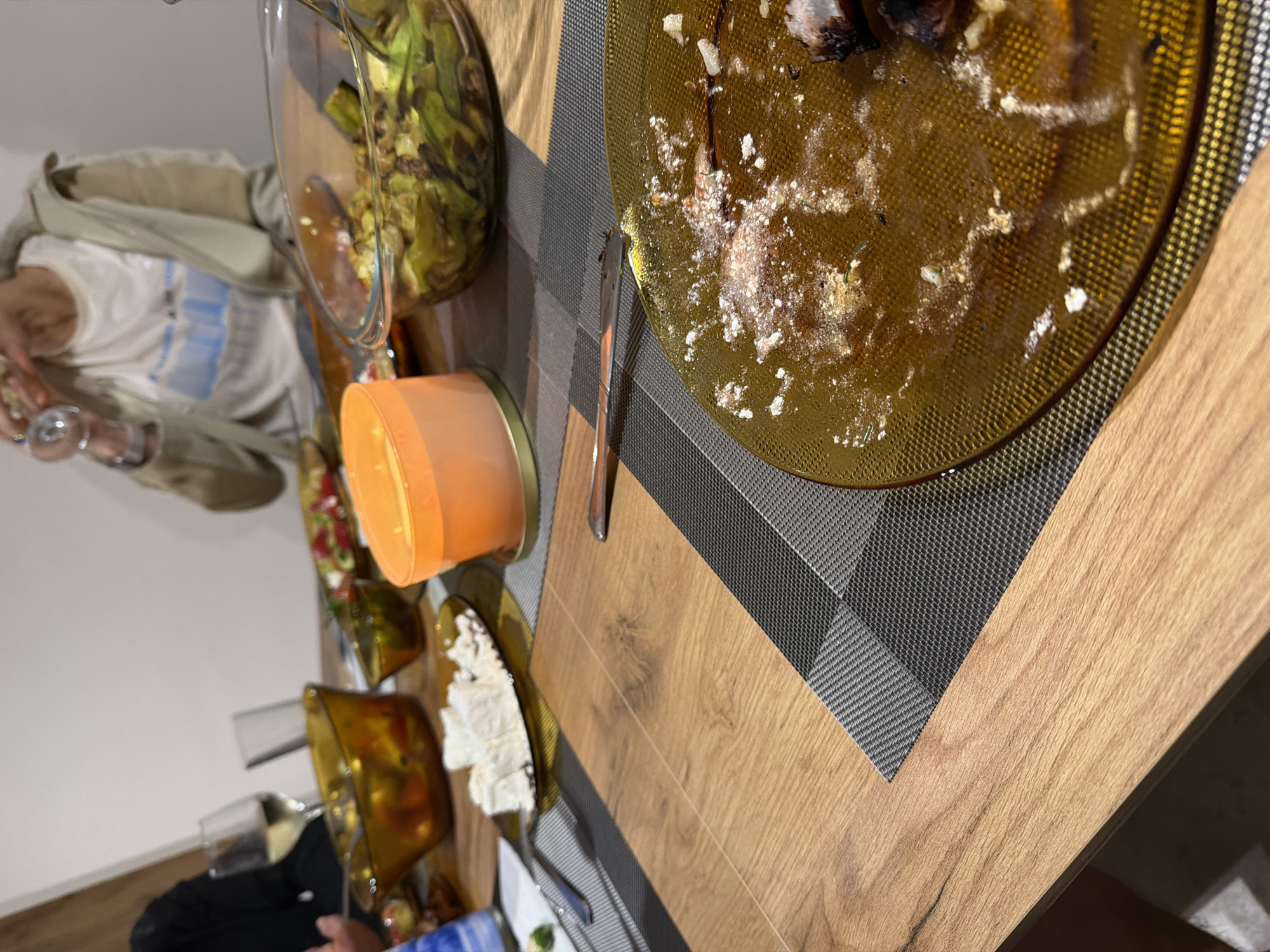 The image shows a table setting with food and drink remains, likely after a meal. A partially eaten plate sits on a grey patterned placemat on a wooden table. There's a salad bowl and possibly other food items nearby, as well as a glass with liquid. A person wearing a tan jacket and a patterned shirt is partially visible. The location, Litakovo, Bulgaria, is assumed but not evident in the image. There is no explicit action or interaction visible other than the inferred consumption of food.