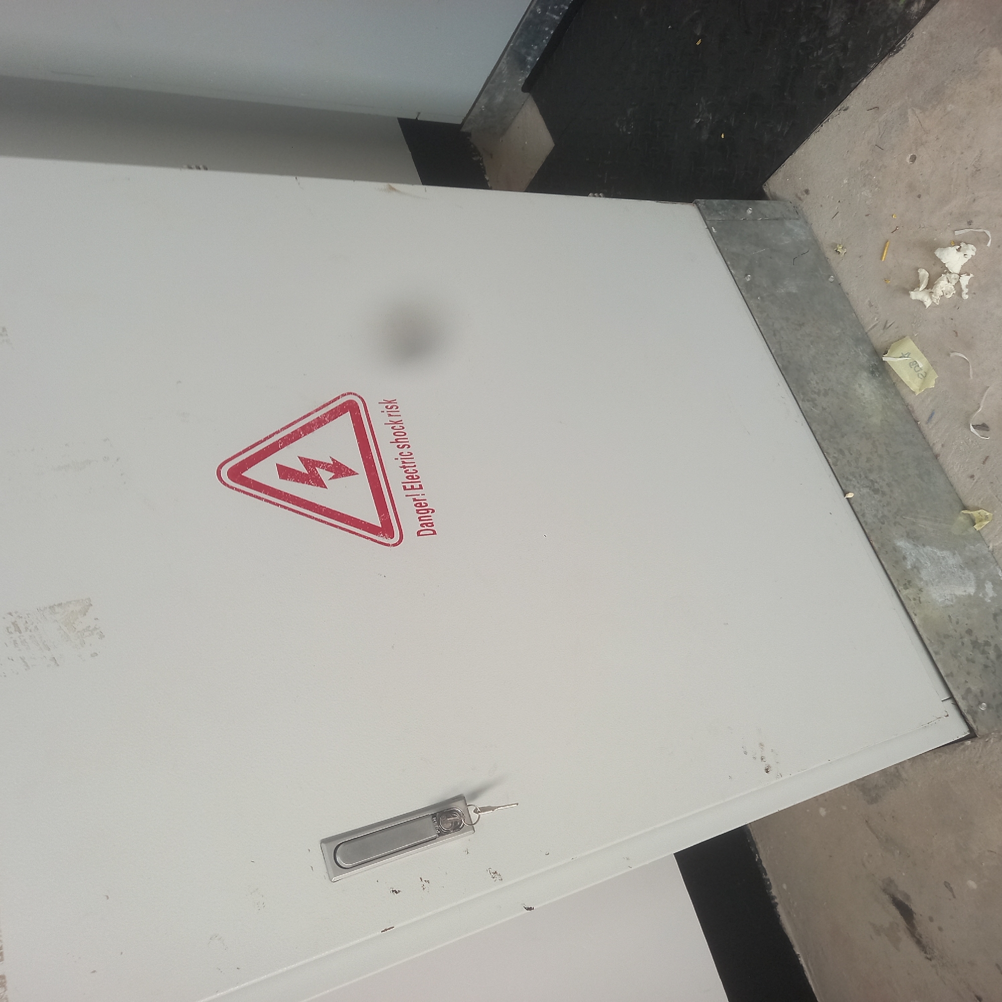The image shows a close-up view of a white electrical cabinet with a warning sticker. The sticker is red and triangular, with a lightning bolt symbol inside, and the text "Danger! Electric shock risk" below it. The cabinet has a silver handle with a key inserted. The cabinet is located in a dimly lit room with a concrete floor. The floor is dirty and has some debris on it, including paper scraps. The overall impression is one of an industrial or utility area.

The location is described as being in the city of Bububu, Tanzania. However, the visual evidence within the image does not provide any specific details that would confirm or deny this location. The environment appears to be indoors, with an electrical cabinet, concrete flooring, and a black floor covering in the background. There are no distinctive architectural features or cultural markers visible that would pinpoint it to Bububu or Tanzania. The scene is static and shows no people or animals. The lighting is artificial and somewhat dim. The image captures a corner of the electrical cabinet and the surrounding floor area. The cabinet is a functional piece of equipment, and the warning sticker indicates a potential hazard. The presence of debris suggests that the area may not be regularly cleaned or maintained.