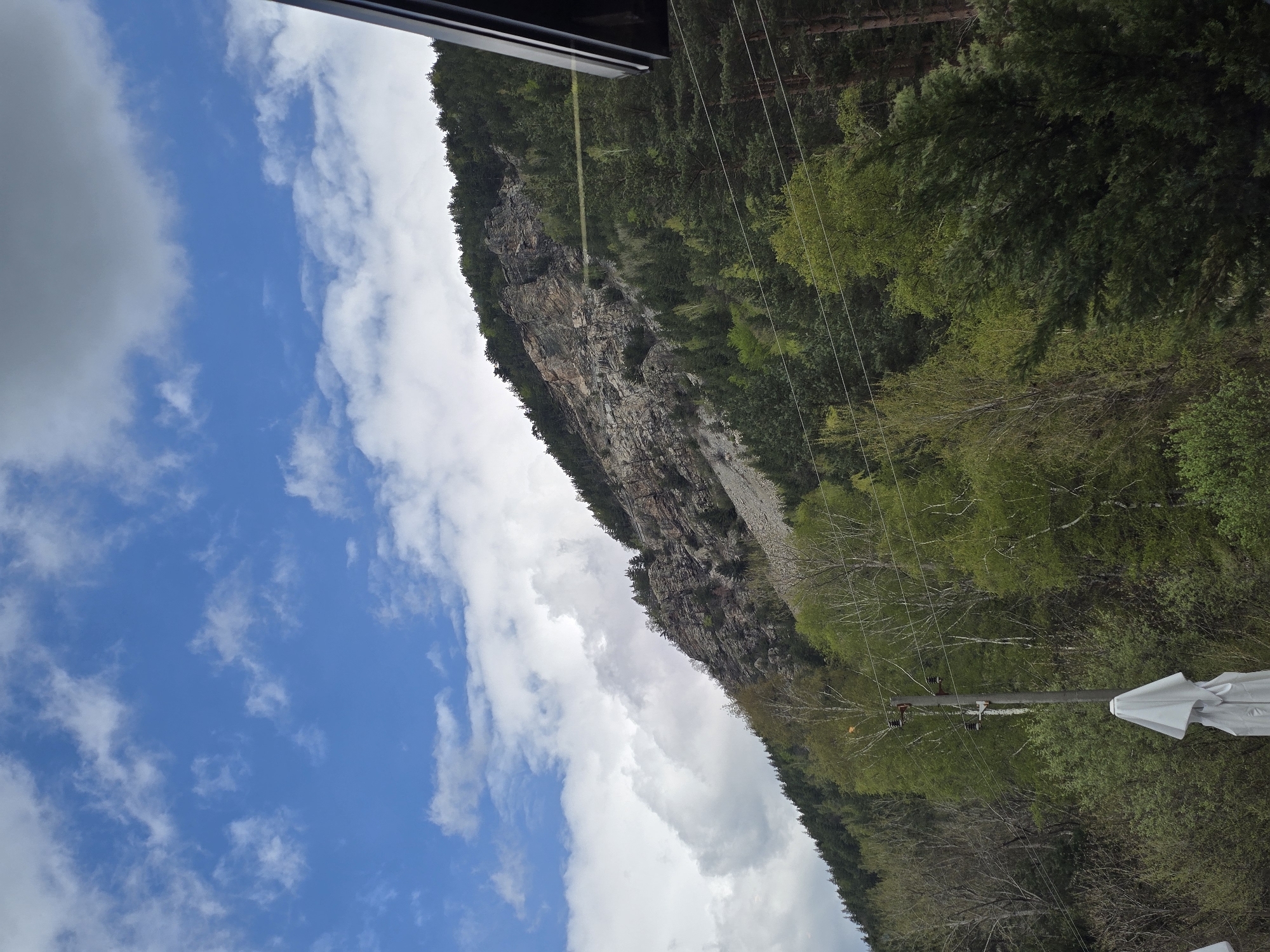 The image shows a view of a rocky mountain ridge covered in trees, taken from what appears to be a building in Sapareva Banya, Bulgaria. The sky is blue with white clouds, and the top of the image shows the edge of a window frame. A power line and a portion of a white umbrella are visible in the lower right. The mountain is situated to the right of the sky.