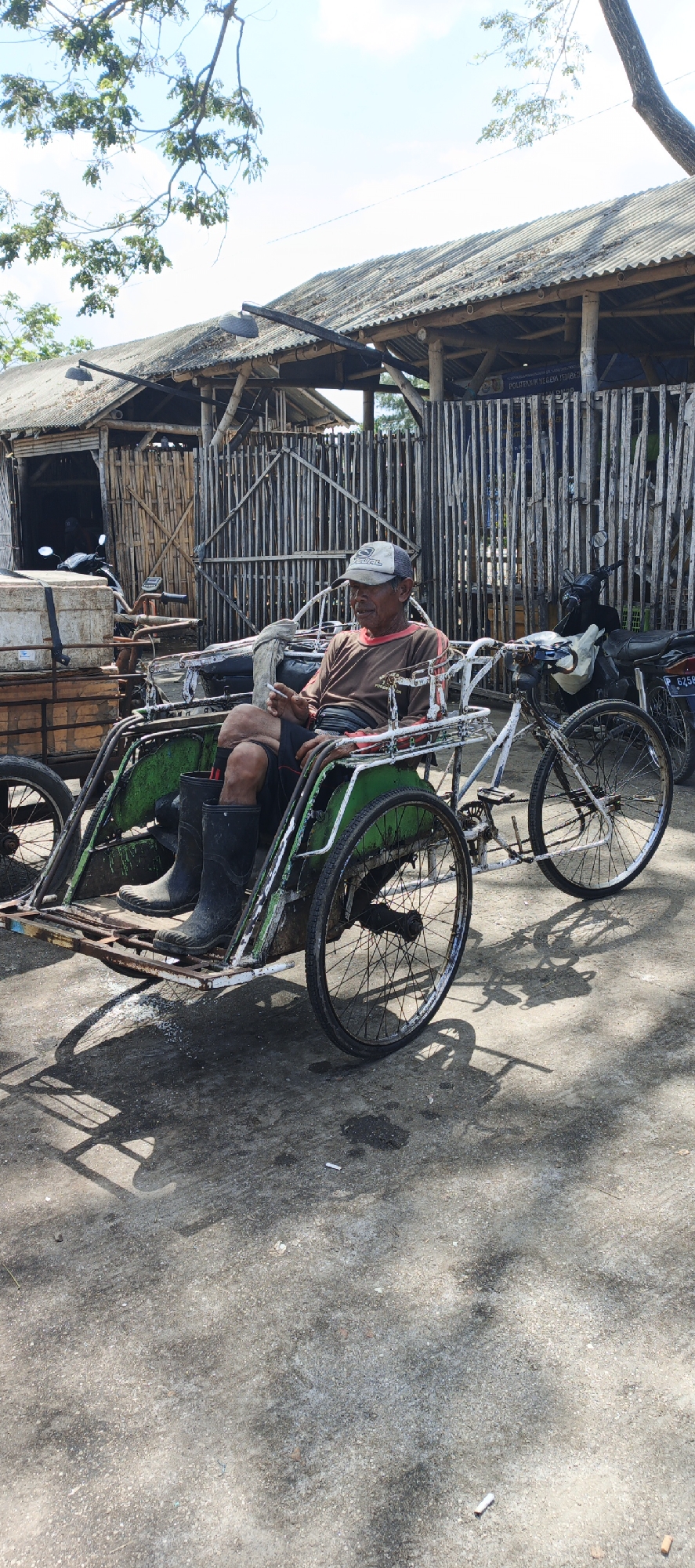 A man wearing a brown long-sleeved shirt, black shorts, a baseball cap, and black rubber boots sits in the driver's seat of a three-wheeled vehicle. The vehicle has a green body, a white frame, and a bicycle wheel at the front and two at the rear. The man is holding a cigarette in his right hand. He is positioned in front of a wooden fence and a rustic structure with a corrugated metal roof. A motorcycle is parked behind the fence. The ground is paved with asphalt. The scene appears to be an outdoor setting in the city of Puger, Indonesia.