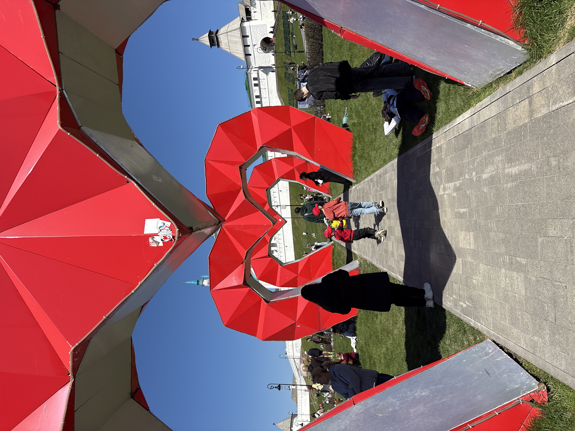 The image captures a public space in Kazan, Russia, showcasing a contemporary red geometric art installation resembling two intertwined figures or an "8" shape. People of diverse ages stand, walk, and sit around the grassy area adjacent to a paved path. The background features a white building with a prominent watchtower-like structure. The day appears sunny, indicated by clear blue skies and distinct shadows cast by the art installation and people. The overall scene conveys a cultural attraction within a historical or urban environment.