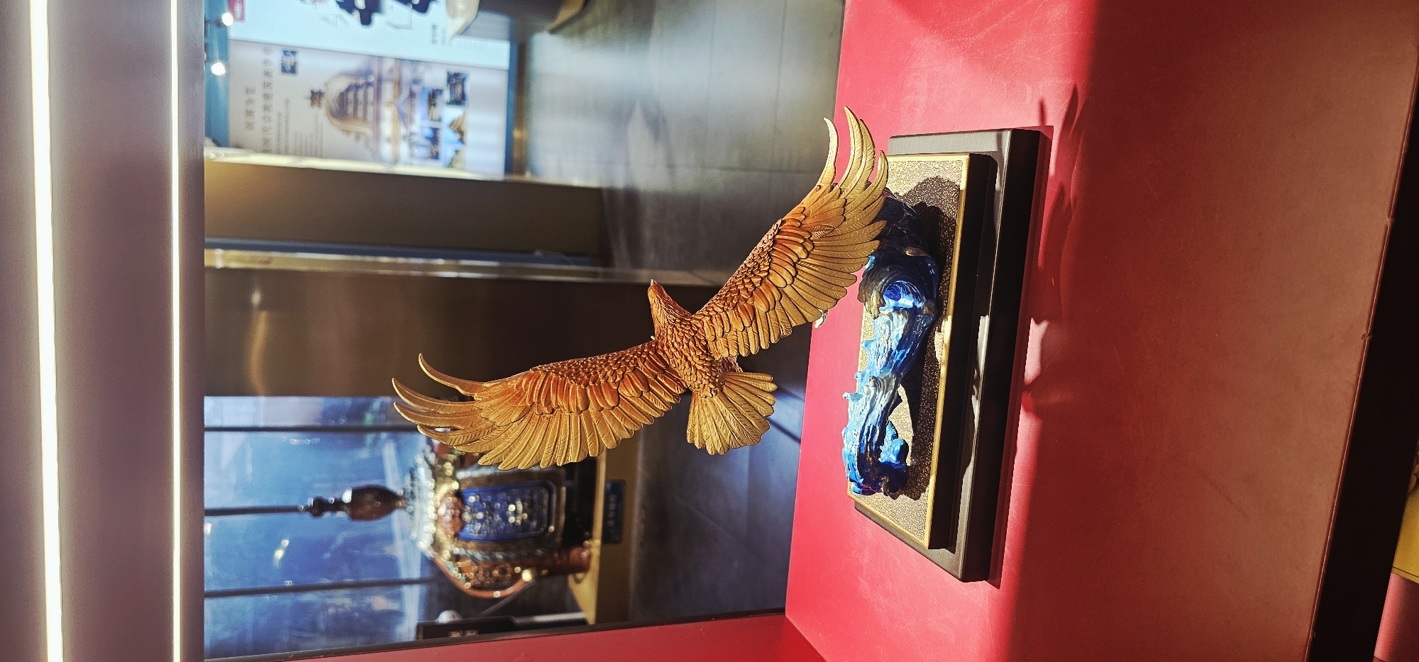 This is a medium shot, eye-level photograph of a golden eagle sculpture displayed on a red surface. The sculpture depicts a golden eagle in mid-flight with its wings outstretched. The eagle is crafted with intricate feather details and has a reddish-gold metallic finish. It is mounted on a dark rectangular base with a textured surface, which in turn rests on a dark wooden platform. Beneath the eagle's wings, a blue, wave-like structure, possibly representing water or wind, is integrated into the base.
The sculpture is positioned in what appears to be an indoor exhibition space or gallery. In the background, through a reflective glass partition, another exhibit is partially visible. This exhibit features a large, ornate vessel with intricate blue and gold detailing, sitting on a display stand. Further in the background, there are large posters or displays with Chinese text and imagery, suggesting a cultural or artistic exhibition. The lighting in the space is soft and appears to come from above, casting subtle shadows.
The photograph captures the sculpture from the right side, with the eagle's head turned to the left. The red surface it rests on is slightly textured and shows some subtle reflections. The overall scene conveys a sense of artistry and display, possibly within a museum or a high-end shop in Yuzhong District, China, a location known for its cultural and commercial significance. There are no people visible in the frame.