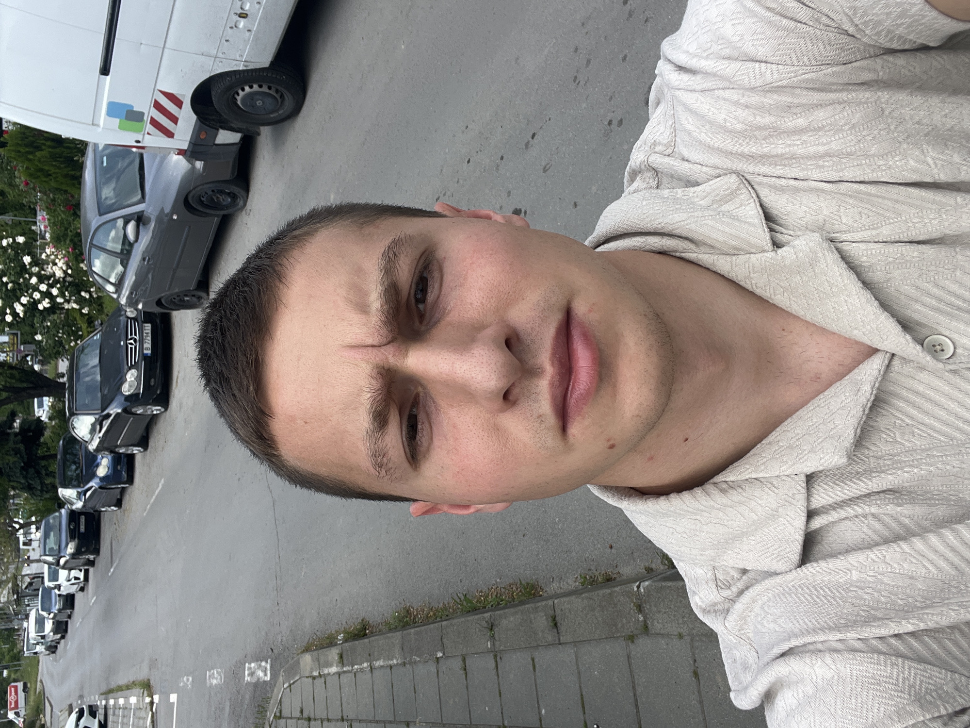 A medium-shot selfie of a fair-skinned, young man with short, dark hair and an angular face. He has a faint scar on his forehead. He is wearing a light-colored, textured shirt with a buttoned collar. The photo appears to be taken outdoors, with a street and parked cars visible in the background. The cars are lined up along the side of the road. A larger white vehicle is partially visible in the top left corner. The image is taken at an angle, as if the phone was held slightly above the subject's eye level. The sidewalk runs along the street with some vegetation. The scene suggests a street in a residential or commercial area with parked vehicles. The cars are of various models, including a Mercedes-Benz. Based on the presence of Mercedes-Benz vehicles and the general urban environment, it can be placed in Varna, Bulgaria.
