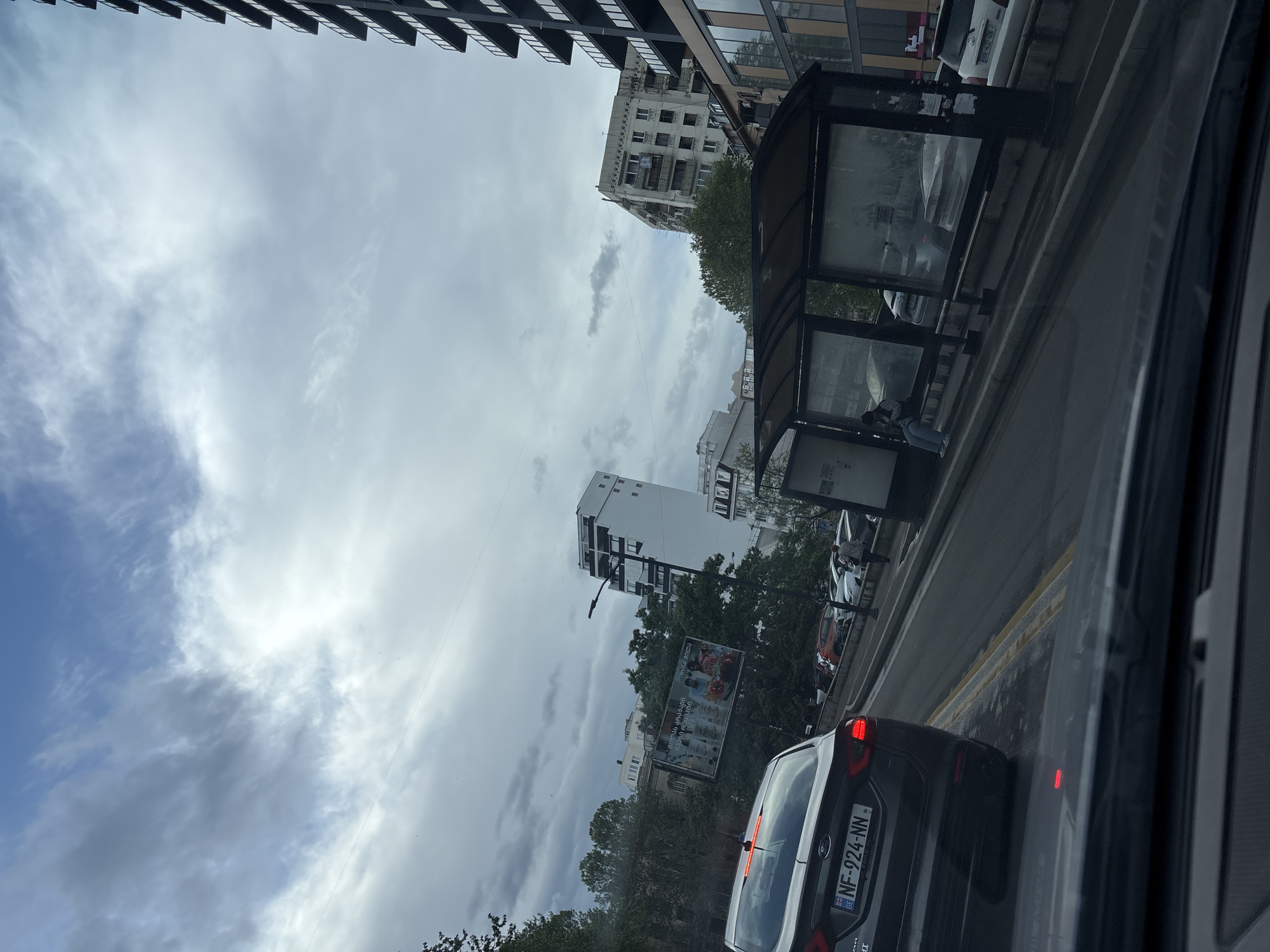 The image, taken from inside a vehicle, shows a street scene in Tbilisi, Georgia. The sky is overcast with a mix of blue and gray clouds. Buildings of varying heights and architectural styles line the street. There is a bus stop structure on the right side of the road. A dark-colored car with a Georgian license plate reading "NF-924-NN" is in front of the vehicle from which the picture was taken. Several other cars can be seen further down the street. Trees are interspersed along the roadside. A large billboard is visible between the buildings.