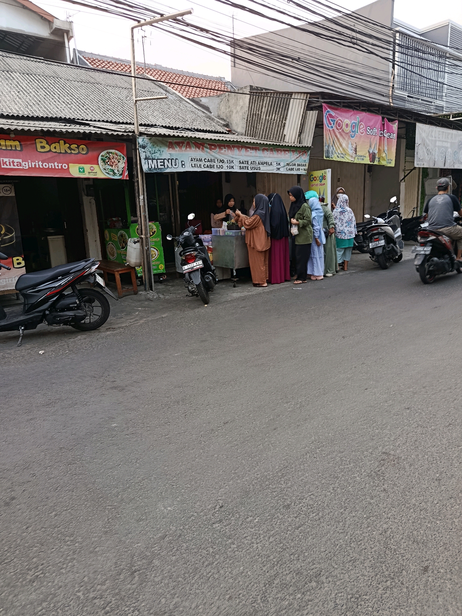In Bekasi, Indonesia, an outdoor scene unfolds along a street lined with small businesses. The foreground is dominated by an asphalt road with several motorcycles parked. A black scooter is parked on the left, and a black motorcycle with a license plate is on the right, further down the street. A man on a motorcycle with a license plate is seen riding away from the camera towards the right.
The middle ground features a row of storefronts. One stall displays a banner that reads "AM BAKSO KITAGIRITONTRO" and shows an image of a bowl of noodles. Adjacent to it, another stall has a banner advertising "AYAM PENYET" and a menu. A line of women dressed in hijabs are waiting to be served at one of these stalls, some interacting with a person behind the counter. Another banner with "Google Soft Ice Cream" is visible further down.
The background shows buildings with corrugated metal roofs and modern structures, suggesting a mix of older and newer architecture. Electrical wires crisscross above the scene, typical of urban Indonesian streetscapes. The lighting indicates it is daytime, with a soft, diffused light. The overall impression is a bustling street-side food stall area in a residential or commercial part of Bekasi.