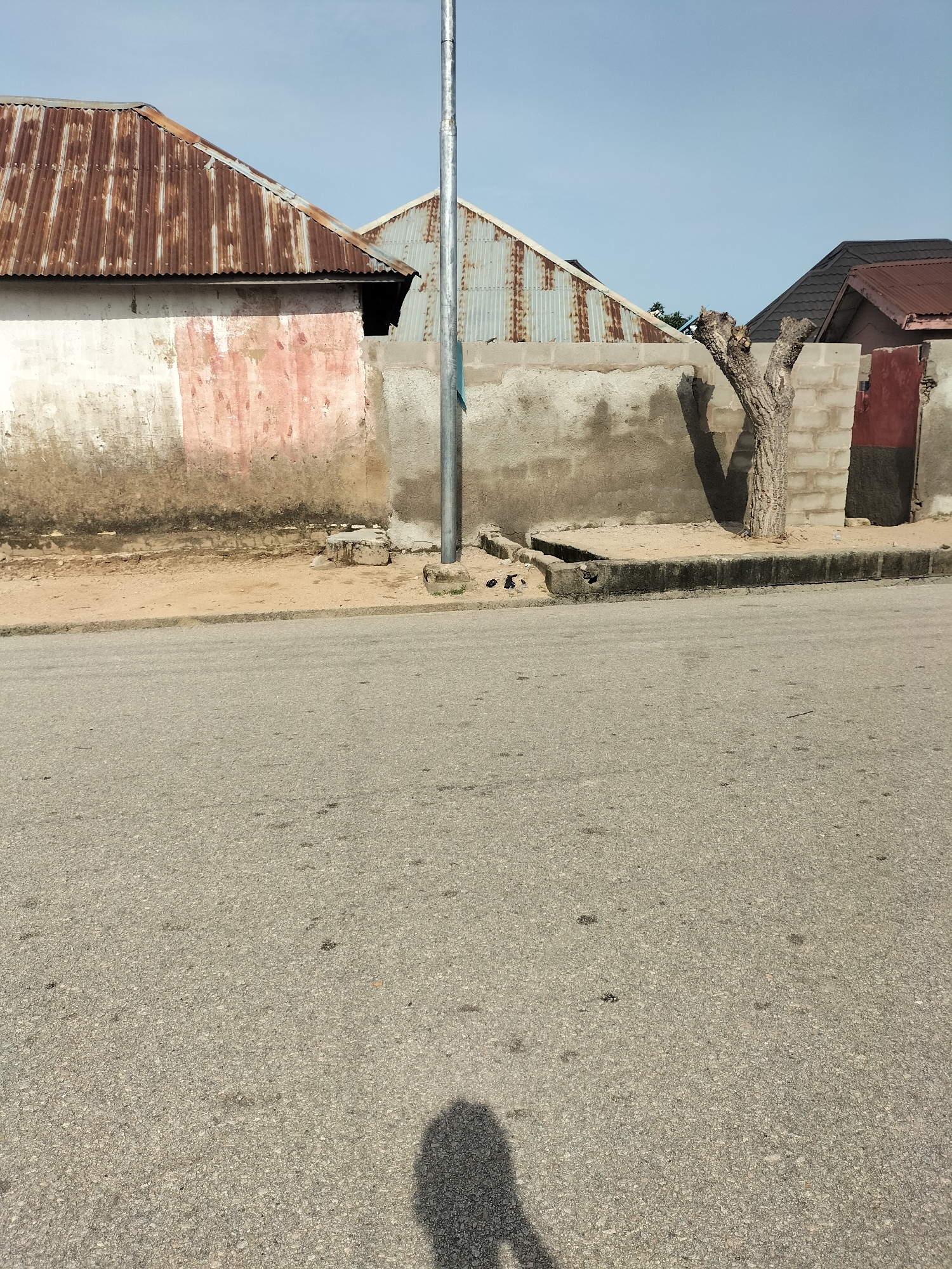 The image depicts a street scene in Jalingo, Nigeria. In the foreground, a grey asphalt road is visible, with a shadow of a person casting onto it from the bottom right. Beyond the road, a low, grey concrete wall runs horizontally across the middle of the frame. Behind the wall, several structures are partially visible. On the left, a building with a worn, pink and white plastered wall has a rusty corrugated metal roof. To its right, another building with a similar rusty corrugated metal roof is situated. Further to the right, a bare tree trunk stands next to a section of brick wall. Behind this, parts of buildings with dark brown tiled roofs are visible. A silver pole, possibly a street light, stands in front of the wall, to the left of the tree. The environment appears dry and dusty, with sparse vegetation visible behind the buildings. The sky is a clear, pale blue.