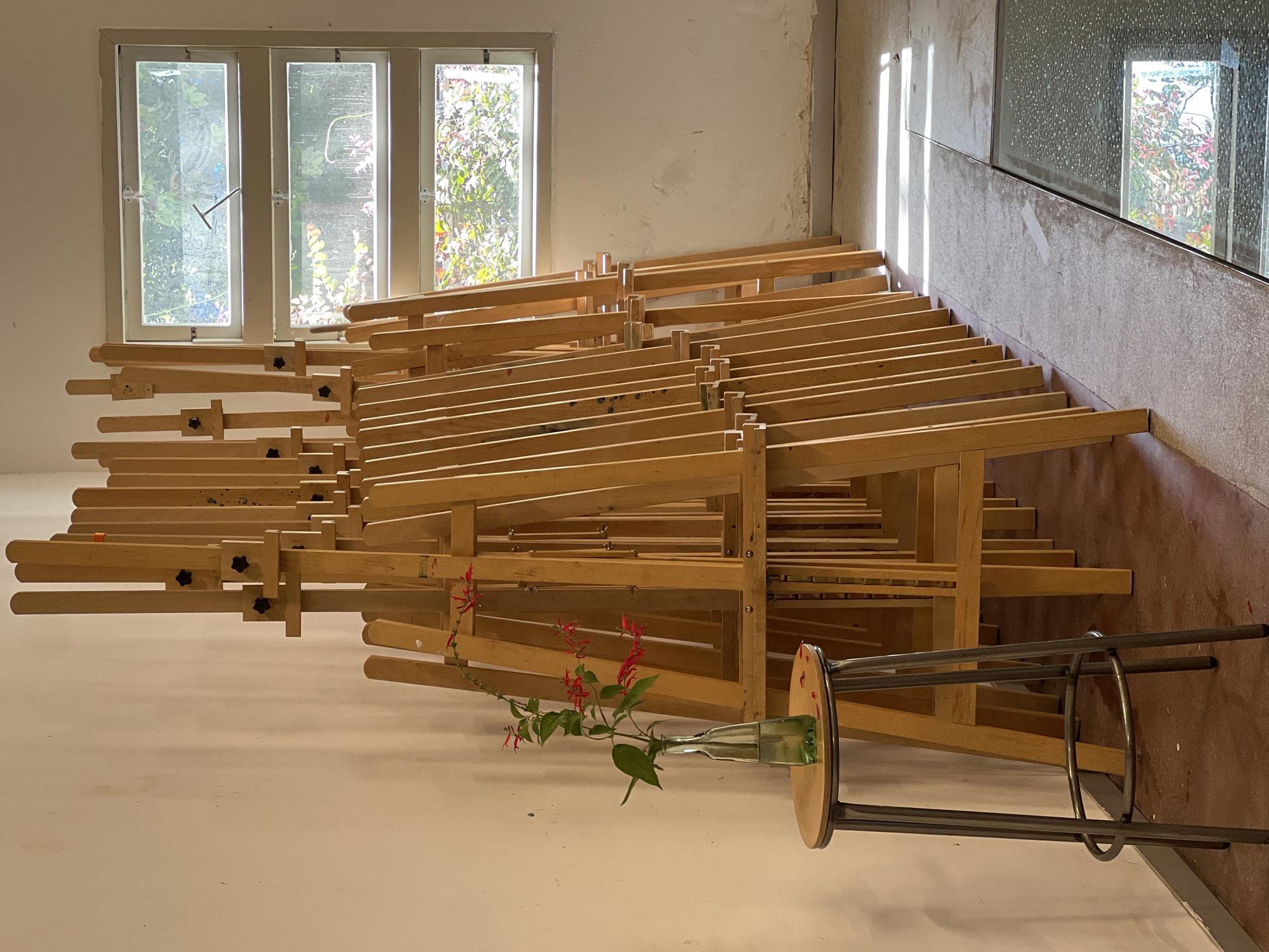 The image shows a room, possibly an art studio, with a large stack of wooden easels filling the foreground. Natural light streams in through a window on the left, illuminating the room and casting shadows. A stool lies on its side with a glass bottle containing red flowers resting on it. The background is partially out of focus, but a second window can be seen in the upper right. This image does not contain enough detail to establish that it is in the city of Tel-Aviv, Israel. More information or details would need to be visible in the photo.