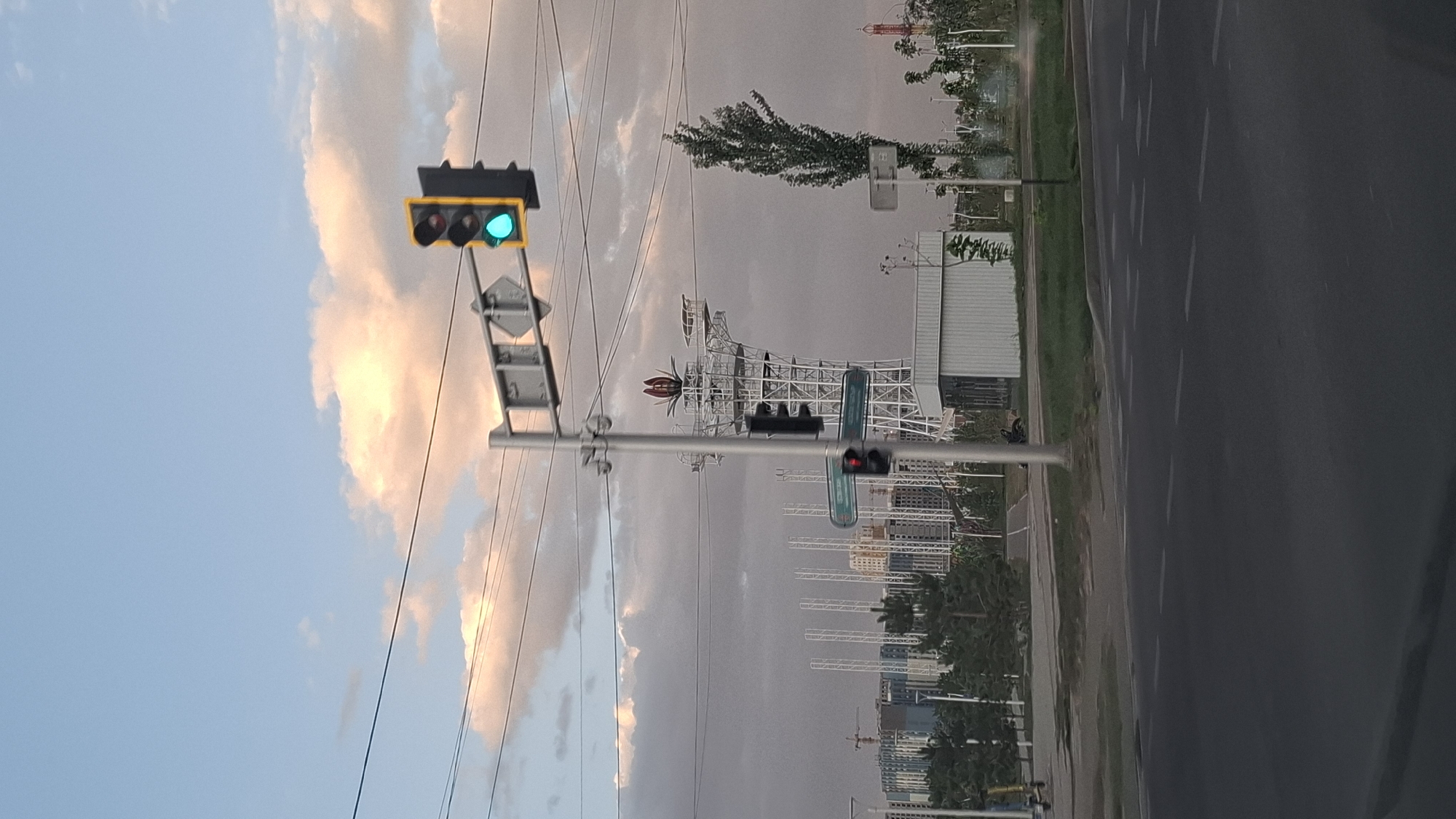 This image captures a street scene in Shymkent, Kazakhstan, likely near the Aqua Park. In the foreground, a road with white lane markings is visible, suggesting it's an intersection. A traffic light is illuminated green, indicating it's safe to proceed. To the left of the traffic light, a tall, white, lattice-like structure resembling a tower with a red flower-like ornament at its peak dominates the background. In front of this structure, there are buildings and trees. Further in the background, construction cranes are visible, indicating ongoing development. A tall, slender tree stands to the right of the tower. The sky is a mix of blue and clouds, with some illuminated by sunlight, suggesting either dawn or dusk. There are power lines stretching across the upper portion of the image. The overall impression is an urban environment with modern architecture and infrastructure, under a cloudy sky.