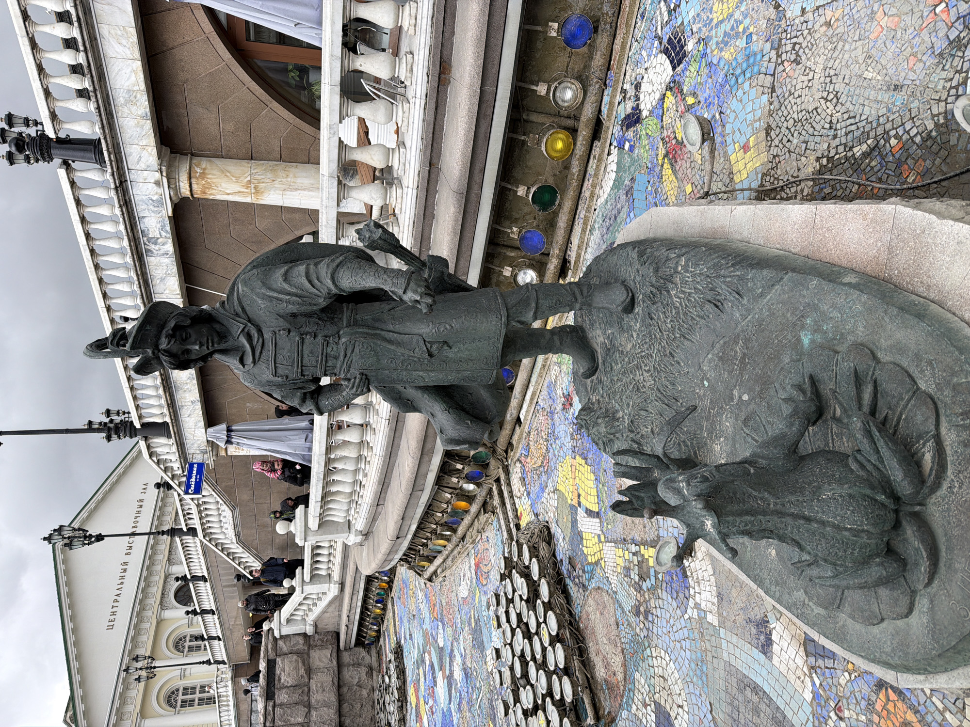 The image captures a scene in Moscow, Russia, featuring a bronze statue of a man standing over a lizard-like creature, likely a depiction of a character from Russian folklore. The statue is placed against a backdrop of colorful mosaic tiles and is positioned near a building identified as the "Central Exhibition Hall," as indicated by signage visible on the building's facade. The architectural style of the building includes classical elements like columns and balustrades. A group of people can be observed on the balcony of the building, suggesting it is accessible to visitors. The weather appears overcast, with a grey sky visible above the building. The statue is detailed, with the man depicted wearing a distinctive hat and clothing style reminiscent of historical attire. The mosaic wall is elaborate, composed of many small pieces of varying colors, creating a patterned effect. Additional architectural elements, such as decorative lighting fixtures, are present along the wall adjacent to the statue.