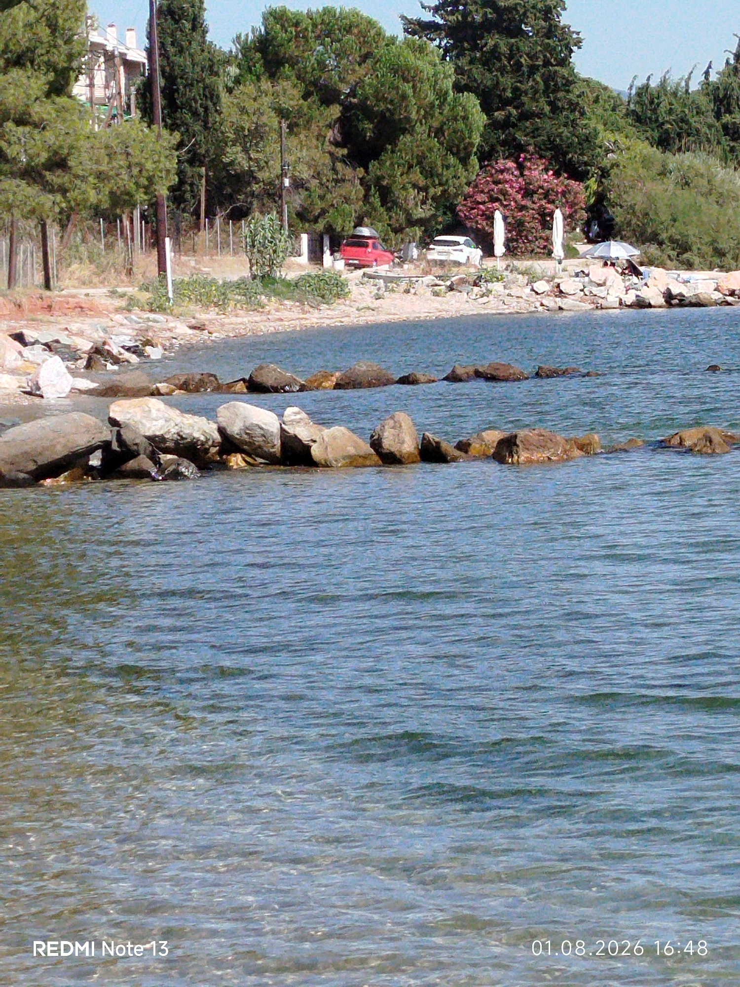 The image shows a rocky beach with the Aegean Sea in the foreground. In the background, there is a dense line of trees, and behind that, some buildings. There are two cars parked near the trees. A few people are visible sitting on the rocks on the right side of the image, some under umbrellas. The water is clear and blue, with small waves. The shoreline is made up of pebbles and rocks. The text on the bottom left corner reads "REDMI Note 13", and on the bottom right corner reads "01.08.2026 16:48". This appears to be a scene from Sithonia Municipal Unit, Greece.