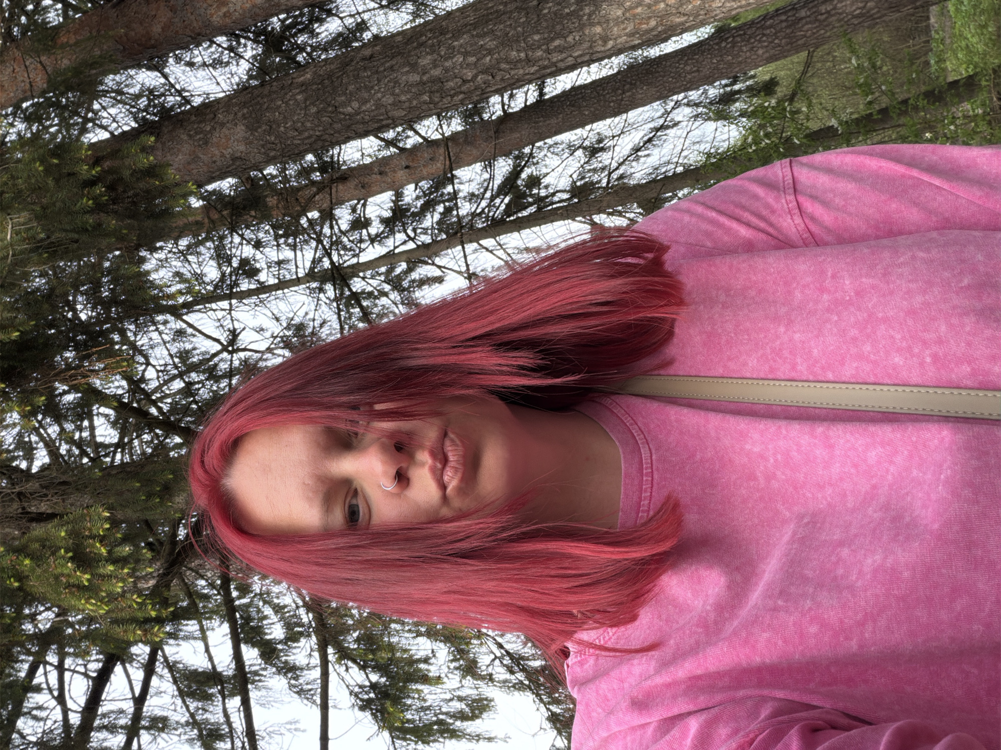 A woman with red hair is wearing a pink sweater and standing in a forest. She has a nose ring. The location is likely in or near the city of Chaikovsky, Russia, given the request. Tall trees are visible in the background. The picture seems to be taken from a low angle, looking up at the subject and the trees.
