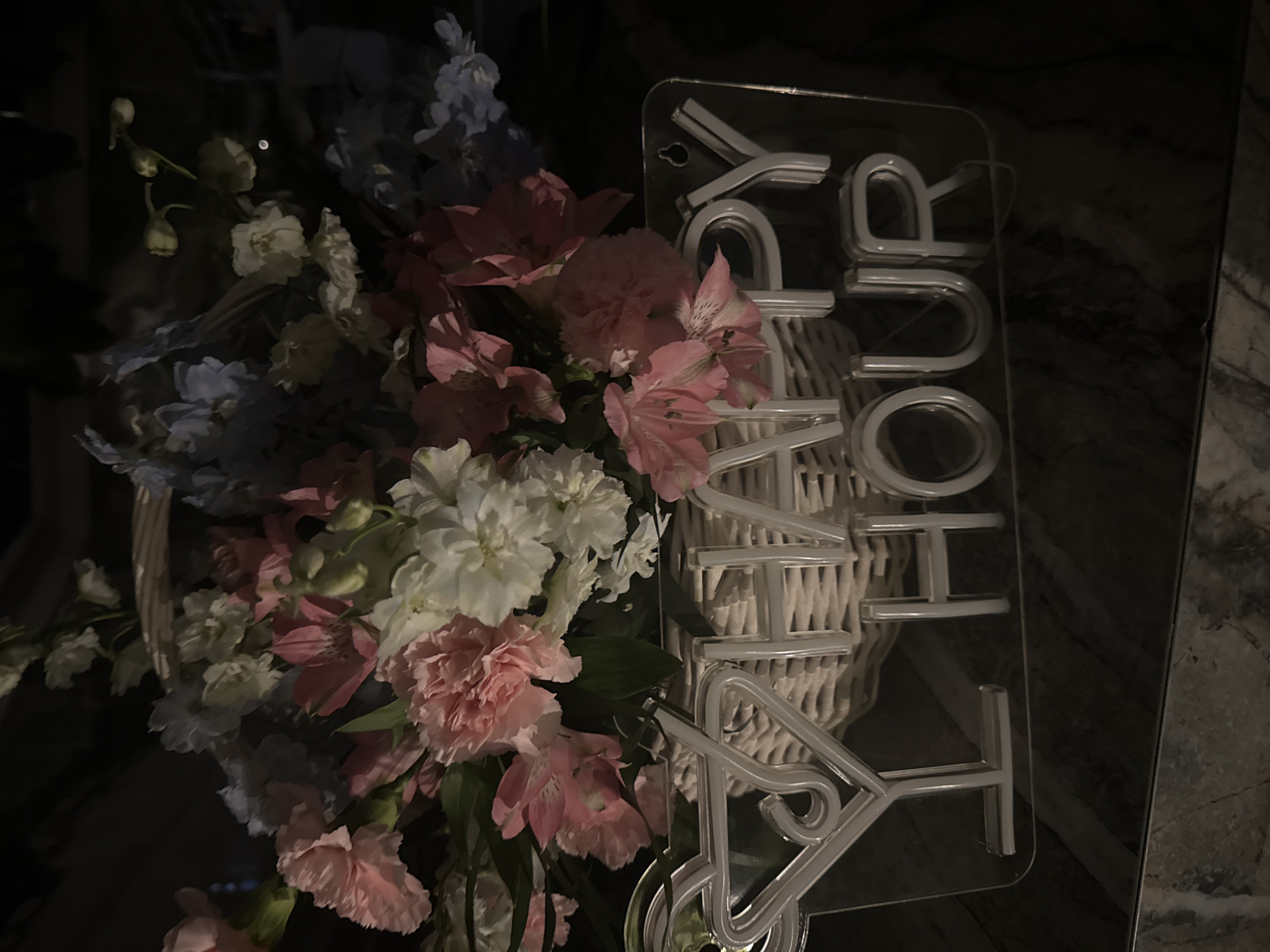 A close-up, low-angle shot captures a woven basket overflowing with a vibrant arrangement of pink, white, and light blue flowers. The flowers appear to be a mix of carnations, alstroemeria, and delphiniums. In front of the basket, a clear acrylic sign with illuminated white neon tubing displays a martini glass icon followed by the words "HAPPY HOUR." The sign is mounted with a small screw at the top right. The background is dark and out of focus, obscuring any specific environmental details. The scene is illuminated by the neon sign and ambient lighting that highlights the flowers and the sign. The image is undated and the specific location within Agios Tychona Community, Cyprus, cannot be determined from the visual information provided.