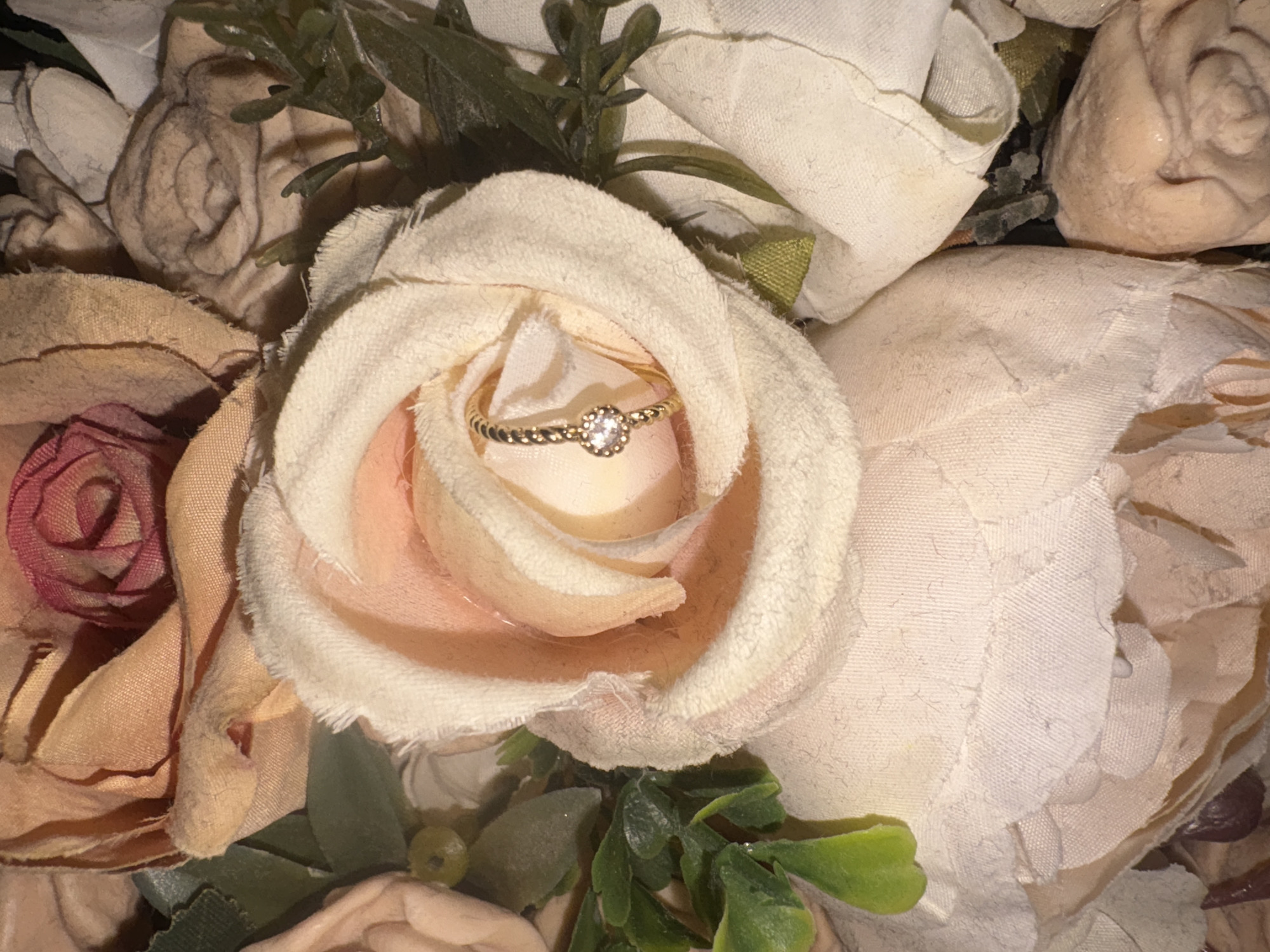 The image shows a close-up of a bouquet of artificial roses in shades of cream, blush, and peach. A small, gold ring with a clear stone sits nestled in the center of a cream-colored rose. The roses are made of fabric, with some petals appearing slightly frayed. Green faux foliage is interspersed among the flowers. There are no people or discernible geographical features visible, therefore no information can be provided about its location in Georgiyevsk, Russia.