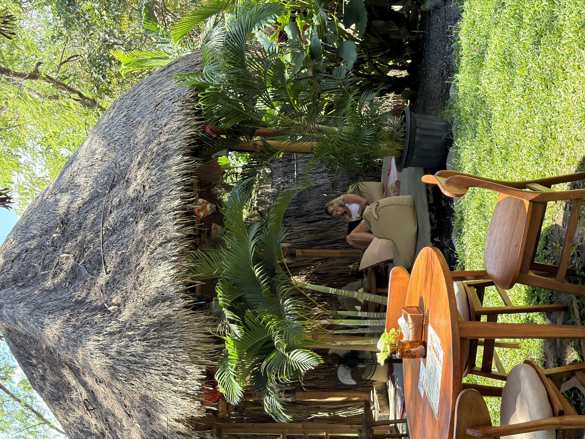The image depicts an outdoor seating area at a cafe or restaurant in a tropical environment, likely in Kediri, Indonesia. In the background, a circular structure with a thatched roof and bamboo walls serves as a covered seating area. Inside this structure, a woman with blonde hair, wearing a white tank top, is seated on a large, tan floor cushion and looking at her phone. She is surrounded by lush greenery, including palm trees and other tropical plants. A black oscillating fan is visible behind a cluster of palm plants in front of the covered structure.
In the foreground, a round, wooden table is set with menus, a small vase containing green foliage, and a tissue box. Three wooden chairs, two with light-colored cushions, surround the table. The ground is covered in vibrant green grass. Sunlight filters through the trees, casting dappled shadows. The overall atmosphere suggests a relaxed, natural, and open-air dining or lounging experience.