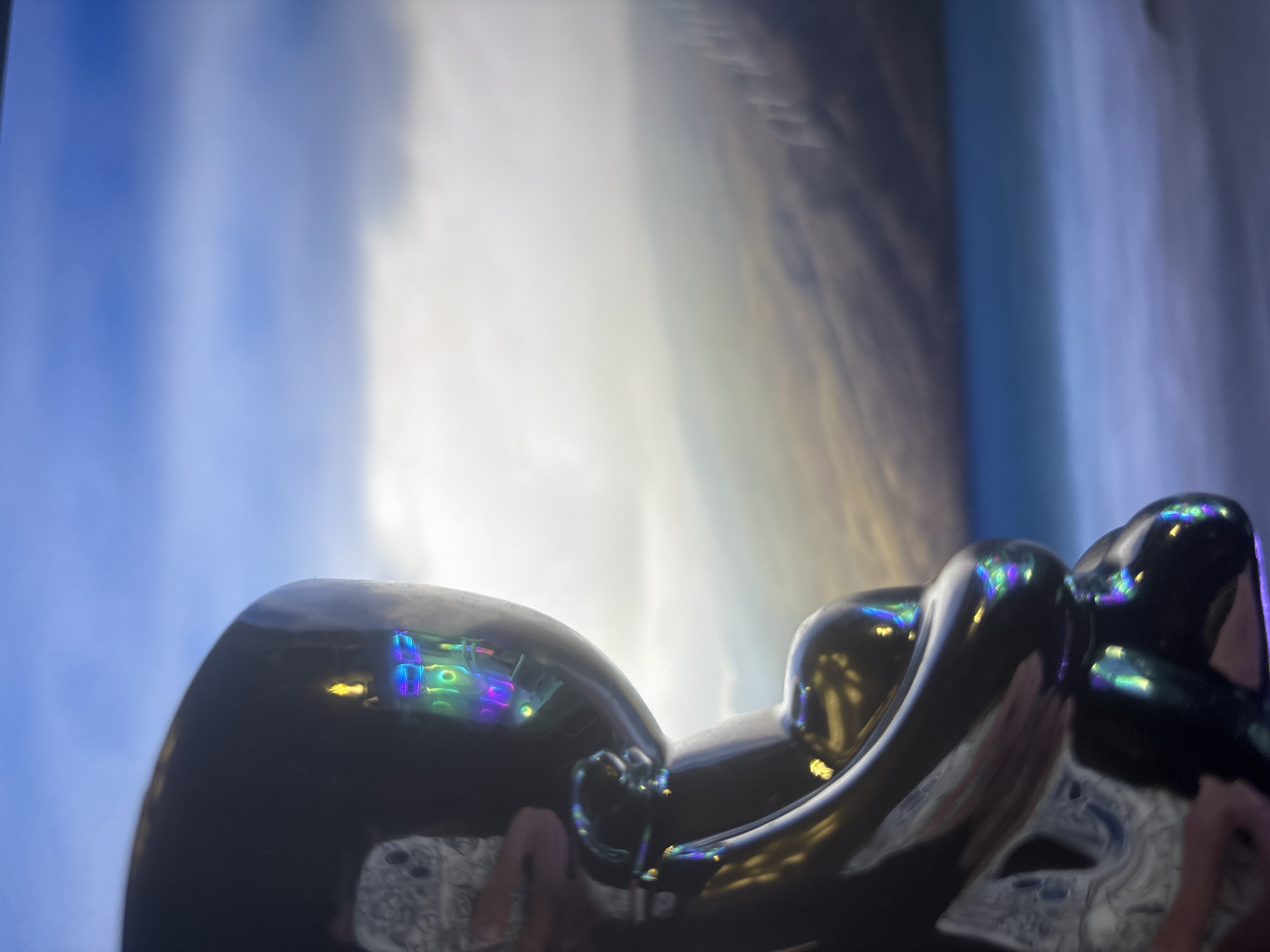 A black, glossy, stylized human figure is positioned in the foreground, facing left. The figure's surface reflects colorful lights, possibly from a screen or other electronic device, showing blues, greens, purples, and yellows. The figure appears to be in a seated or hunched position, with its arms bent and held near its body.
Behind the figure, a blurry landscape unfolds. A body of water, likely a lake or ocean, is visible in the mid-ground, transitioning from a bright turquoise to a lighter blue. Beyond the water, hills or mountains rise, their slopes covered in muted brown and green tones. The sky above is a gradient of blue, with wispy white clouds. The overall impression is of a serene, somewhat abstract scene. The specific location within the city of Unknown, Unknown cannot be determined from the image.