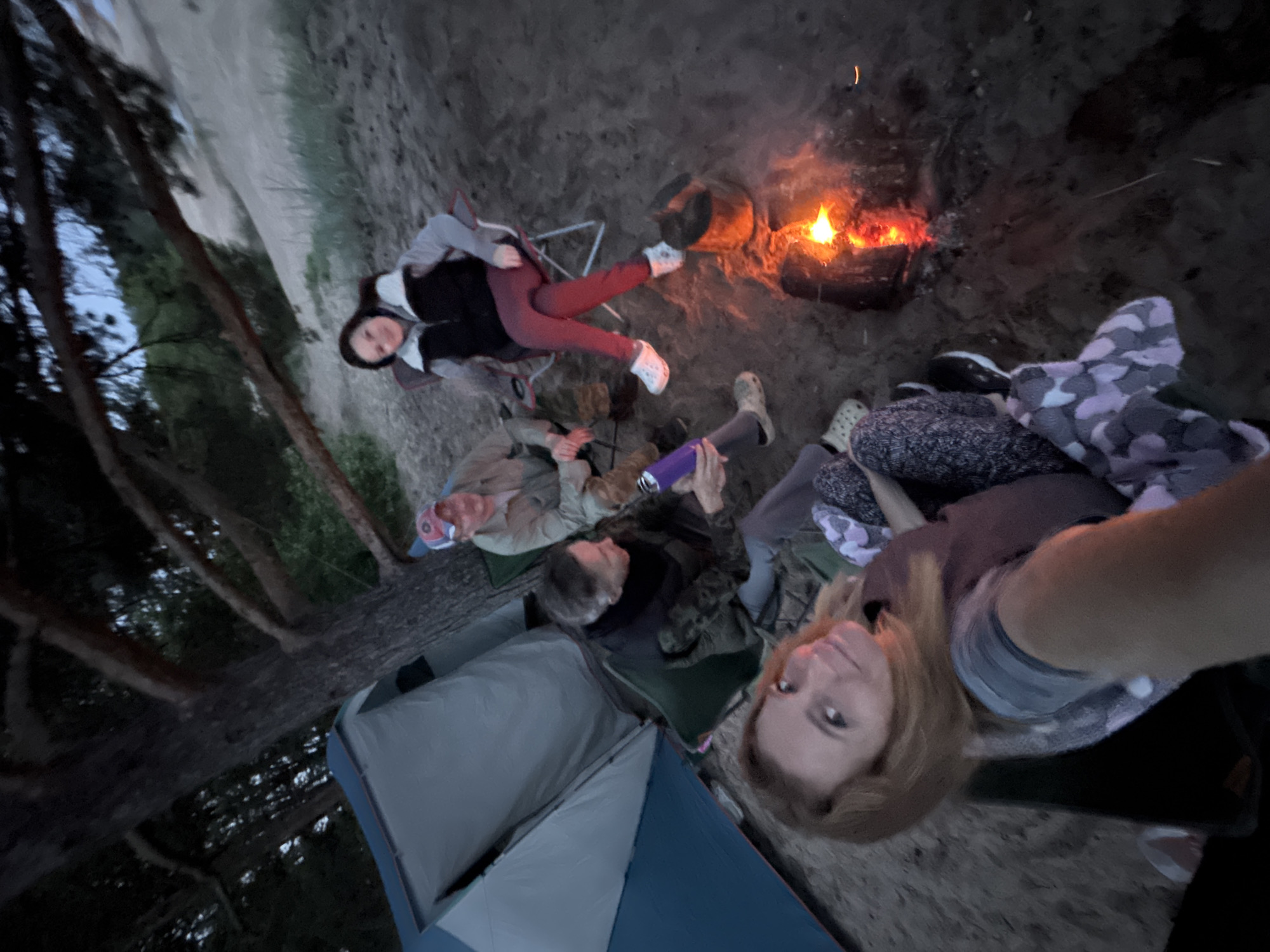 The image depicts four adults engaged in a social gathering around a campfire in a sandy outdoor environment in Unknown, Russia, under conditions of dusk or early night.
On the far left, a two-tone tent, featuring light grey and teal-blue sections, is partially visible, positioned beneath the dark, sprawling branches and trunk of a large tree that occupies the upper left quadrant of the frame. The ground throughout the scene is composed of light-colored sand, characteristic of a beach or dune landscape, with minimal sparse grassy vegetation present on a gentle sandy slope in the background on the right side.
Four individuals are seated in various folding camping chairs arranged around a central campfire. In the immediate foreground, lower left, a female with reddish-blonde hair and fair complexion is looking directly at the camera with a slight smile. She is wearing a grey striped top layered under a darker garment, and a patterned blanket (displaying purple, grey, and white hues) covers her lower body and legs. To her right, a second female is seated in a green folding chair, oriented to her right. She has dark hair, wears a dark neck warmer, a camouflage-patterned jacket or top, and grey pants, with light-colored clogs on her feet. She is holding a purple insulated bottle in her left hand. To her right and slightly behind, a male is seated in a light-colored folding chair, facing left. He is wearing a light-colored long-sleeved shirt or jacket and a red and white baseball cap. On the upper right side of the image, a third female is seated in a red and white folding chair, facing left towards the campfire and the other individuals. She has dark hair, wears a grey long-sleeved top beneath a dark vest, red pants, and white shoes.
A campfire is burning in the lower right portion of the frame, emanating bright orange and red flames from within a small, undefined containment area on the sand. A dark pot or kettle is positioned immediately adjacent to the burning embers. The collective arrangement of the individuals and the campfire suggests a leisure activity.