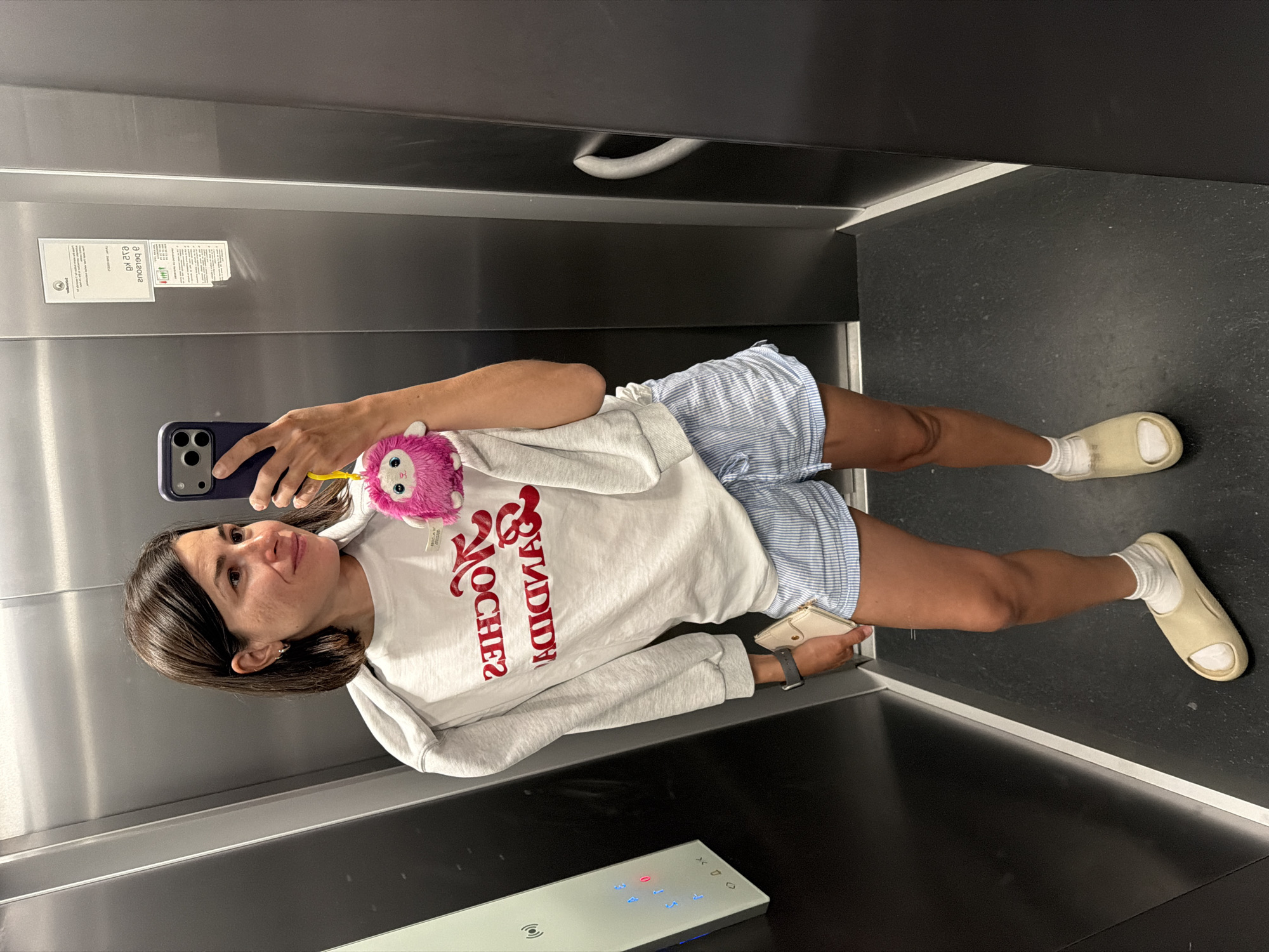 A woman is taking a selfie in an elevator. The woman has brown hair, is wearing a white sweatshirt with red lettering, blue and white striped shorts, white socks, and cream-colored slides. She is holding a blue smartphone with a pink fuzzy object attached. She is also holding a cream-colored wallet. The elevator has stainless steel walls, a black floor, and a control panel with illuminated buttons. There is a sign on the elevator wall with printed text. The location is presumed to be Budva, Montenegro, based on the lettering on the sweatshirt.