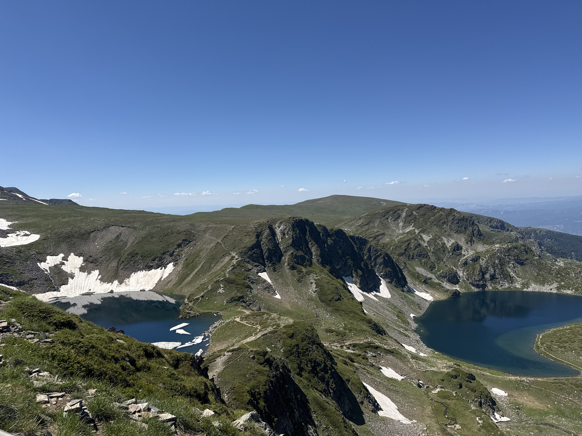 This is a wide-angle, high-altitude view of the Seven Rila Lakes in Rila National Park, Bulgaria, an area often associated with Rila Monastery due to proximity and pilgrimage. The scene captures a clear, bright blue sky above rugged, green mountain terrain. Patches of white snow are scattered across the slopes, indicating a high elevation.
Two distinct glacial lakes are visible. The lake in the foreground and mid-left is a deep, dark blue, with some scattered ice floes on its surface. The lake to the right is larger and also a deep blue, with a lighter, more transparent edge closer to the shore. A winding dirt path is visible, traversing the mountain ridges between and around the lakes, with several small figures of hikers walking along it. The terrain is characterized by steep, rocky inclines covered in low-lying green vegetation and grass. The overall environment is natural, mountainous, and serene, under a cloudless sky.