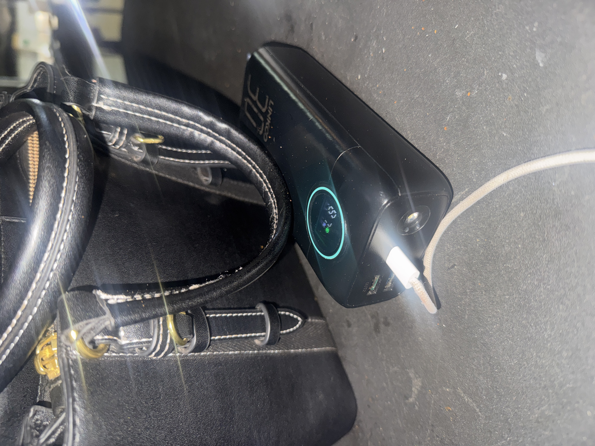 An overhead shot captures a black leather handbag and a black power bank. The handbag, with gold hardware and white stitching, is partially visible in the upper left quadrant of the image. A black USB-C cable is plugged into the power bank, which displays a blue circular indicator with a percentage reading. The scene is set on a dark, textured surface, possibly a table or floor. The lighting suggests an indoor environment, possibly a commercial or domestic setting within Abuja, Nigeria.