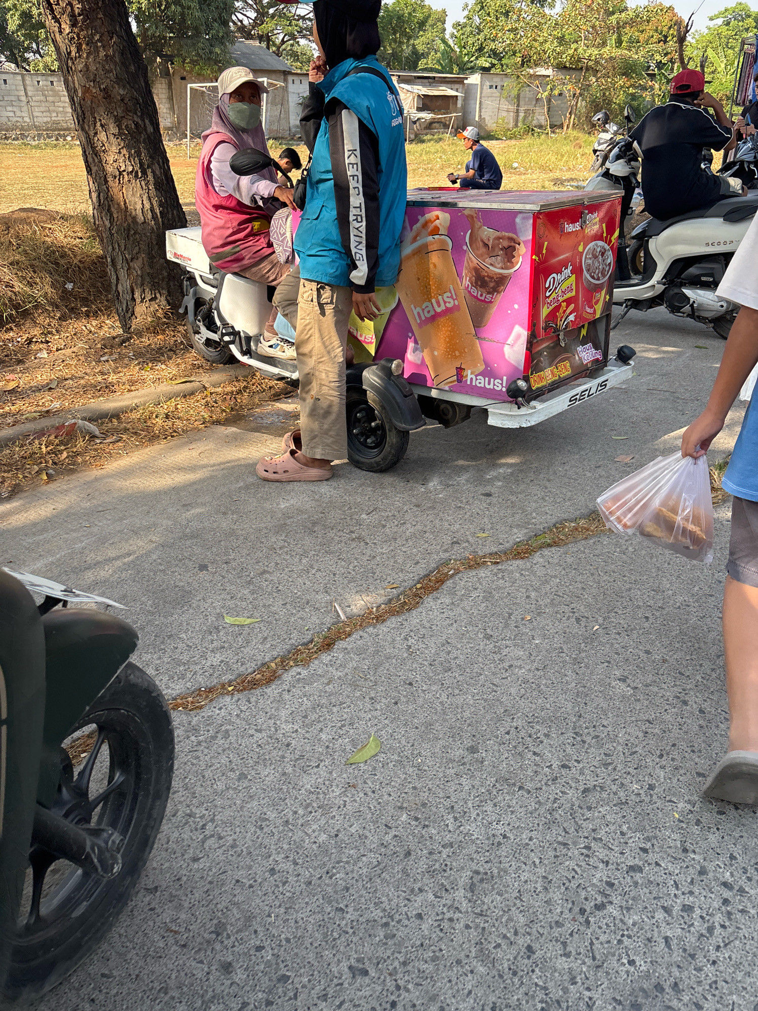 In Telagamurni, Indonesia, a vendor on a trike-cycle cart is preparing to sell cold drinks. The cart, emblazoned with colorful advertisements for "haus!" beverages, is parked beside a busy road. A woman in a pink vest and headscarf sits on the trike, wearing a face mask and light-colored Crocs. A vendor in a blue jacket with "KEEP TRYING" printed on the sleeve stands beside the cart, wearing khaki pants and similar light-colored footwear. In the background, several people are gathered around other vehicles, including a white scooter. A person to the right is holding a plastic bag filled with food items. The scene is bathed in sunlight, casting shadows on the asphalt road.