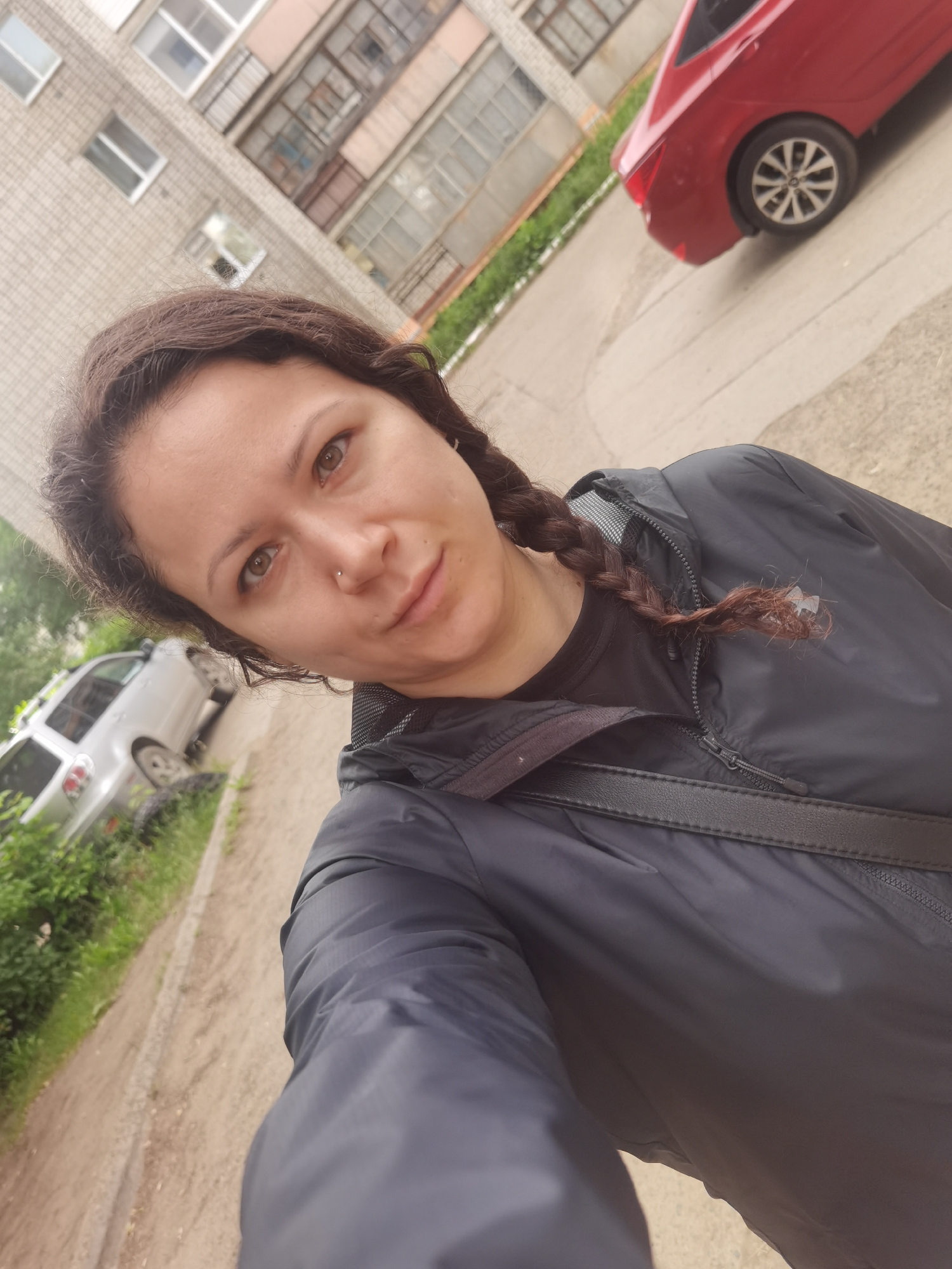 A woman with long, dark brown hair in two braids stands in front of a tiled apartment building in Chaikovsky, Russia. She is wearing a dark, zipped jacket over a dark t-shirt, and a dark strap from a bag is visible across her chest. A red car is parked to her right, and a silver car is parked to her left. The ground is a light-colored dirt or gravel path. The sky is overcast.