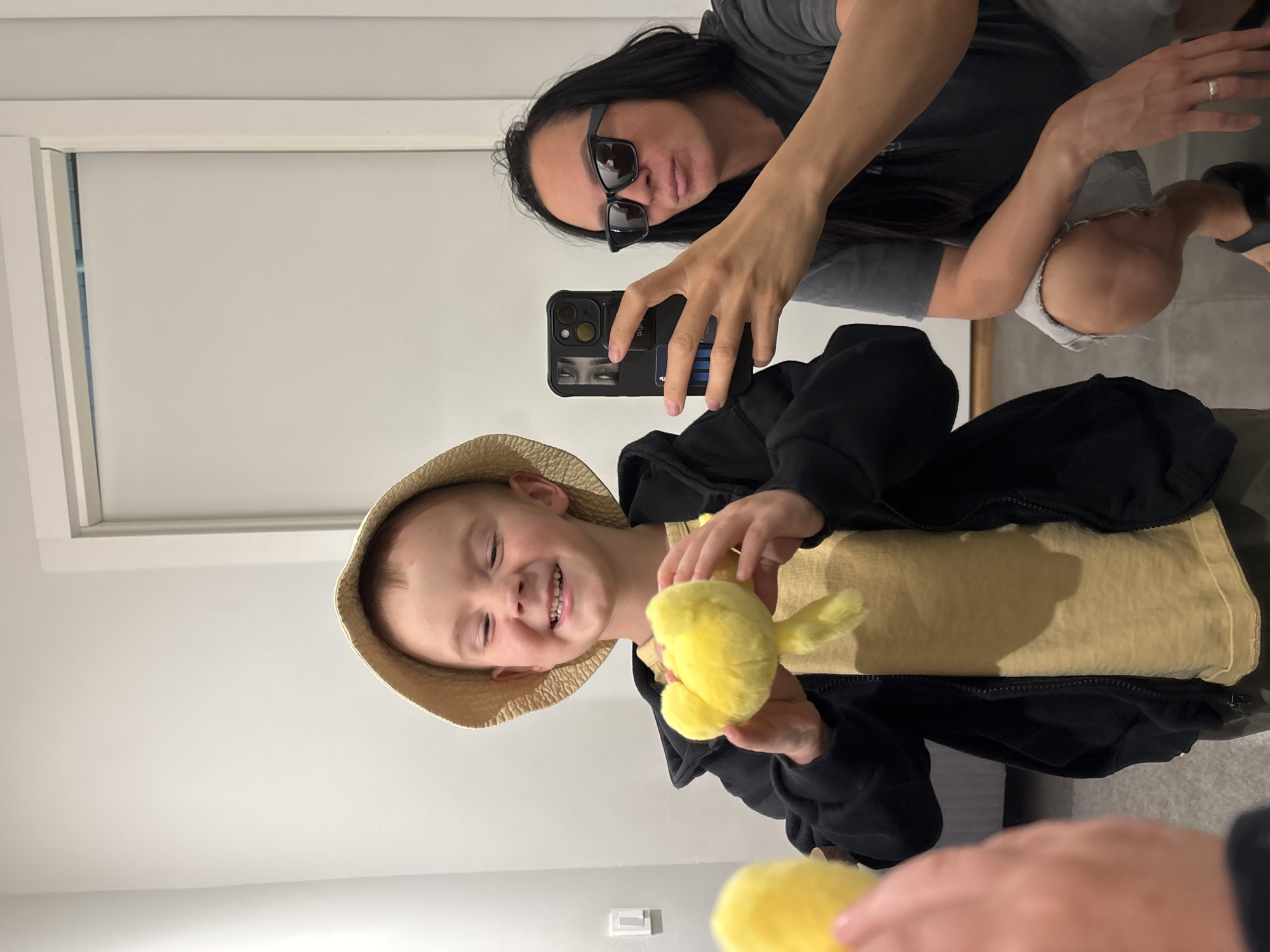 A woman wearing sunglasses is taking a selfie with a young boy in a straw hat. The boy is holding a yellow plush toy, possibly a duck. The photo is taken indoors, with a plain white wall in the background. The city of Chaikovsky, Russia is not apparent from the image.