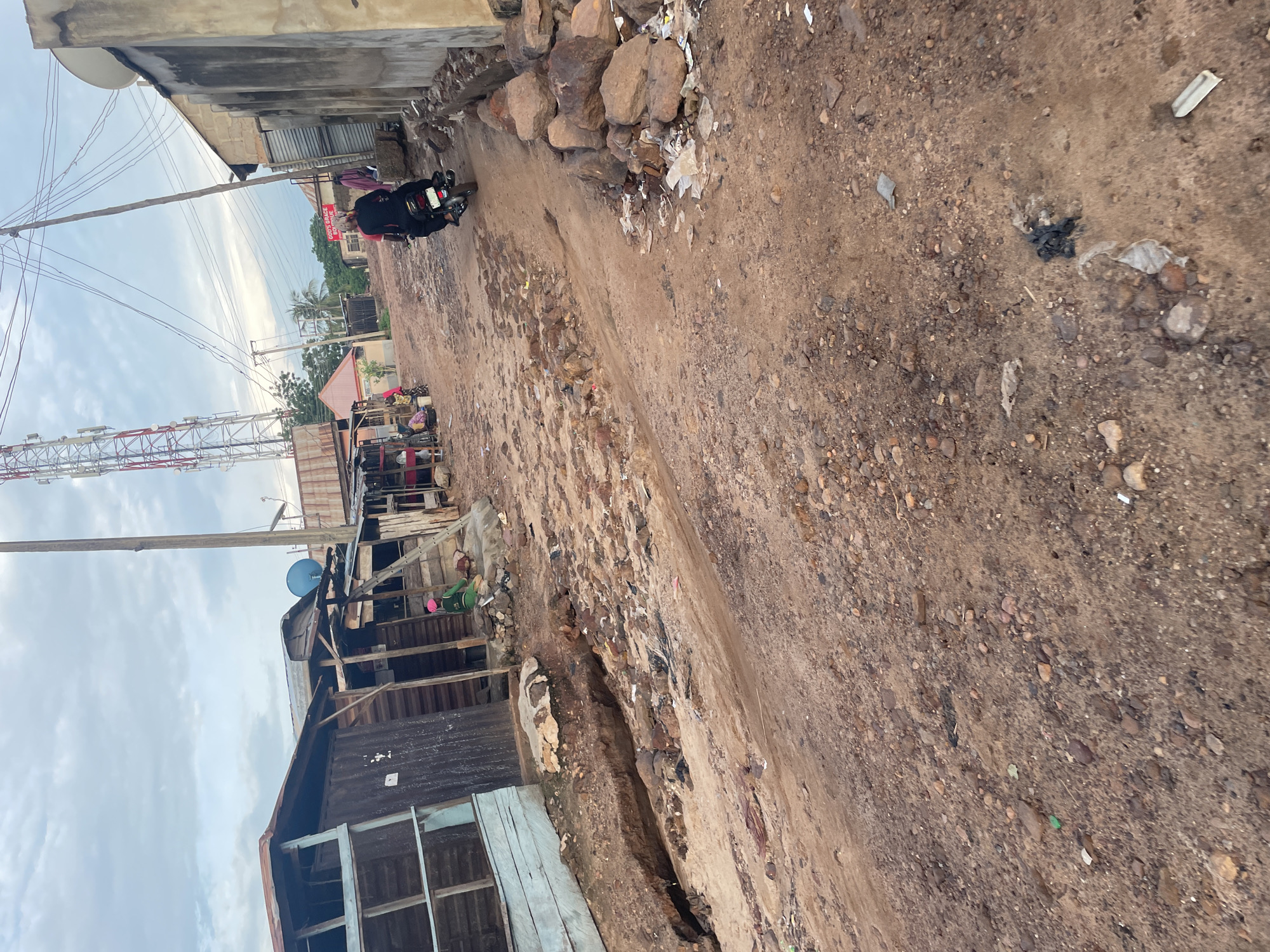 In Lokoja, Nigeria, a dirt road passes through a settlement with makeshift structures. A person on a motorcycle, wearing a black outfit and a headscarf, rides away from the camera. The road is uneven and rocky, with some debris and trash scattered on the ground. On the left, a building with a blue wooden facade and a corrugated metal roof is visible. Further down the road, a telecommunications tower rises against a cloudy sky. The right side of the road features more buildings, some constructed with cinder blocks and others with corrugated metal.