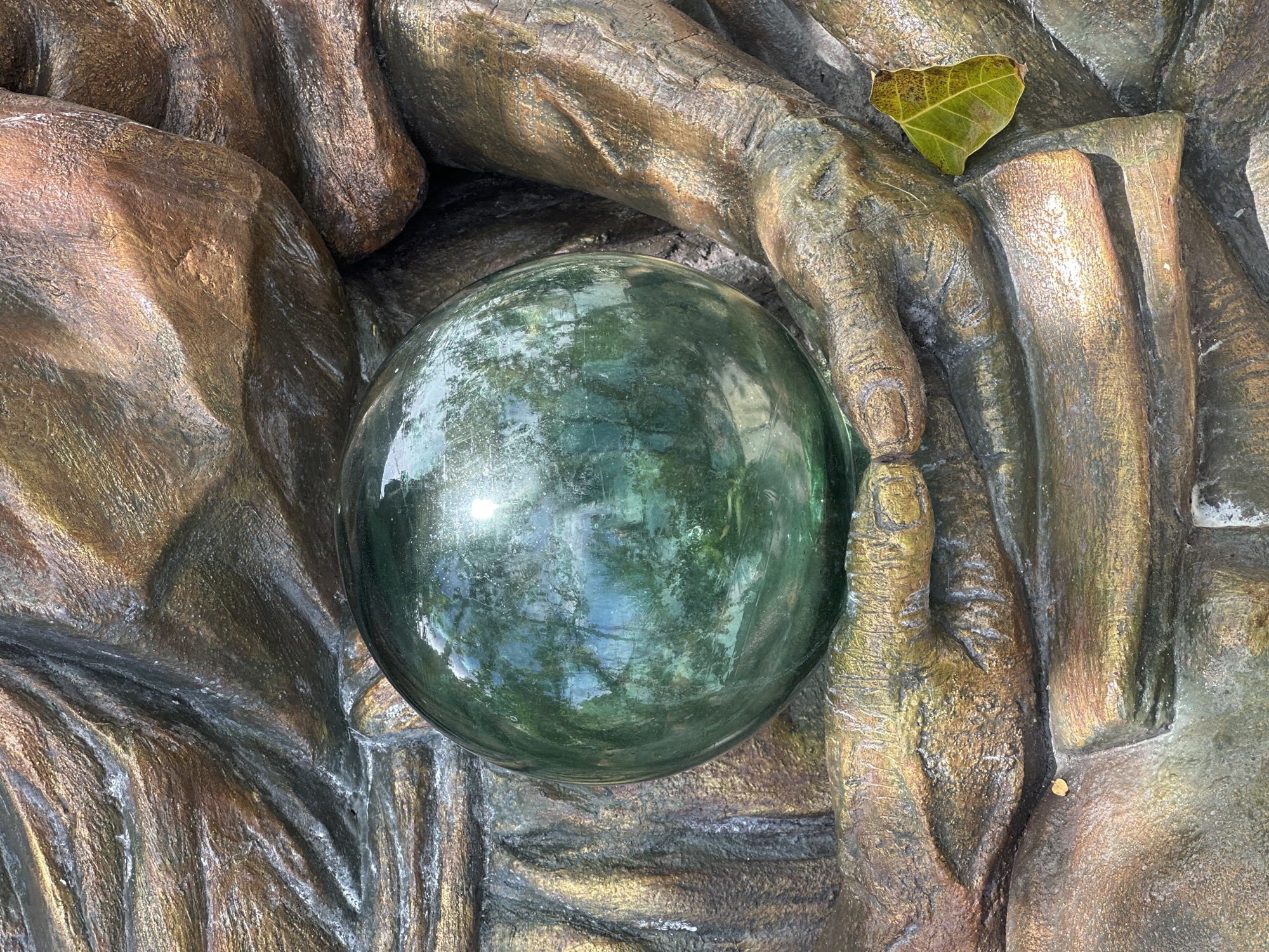 The image shows a close-up view of a green glass orb resting in the cupped hands of a sculpture. The sculpture appears to be made of a material with a bronze-like finish, showing textured folds and creases that resemble fabric or skin. The orb is highly reflective, with the reflections showing trees and sky, suggesting it is outdoors. A single, small, yellow-green leaf rests on the lower right section of the sculpture's hand. The background is filled with the sculpted hands and folds of the artwork. The scene is located in the Koh Phangan subdistrict of Thailand.