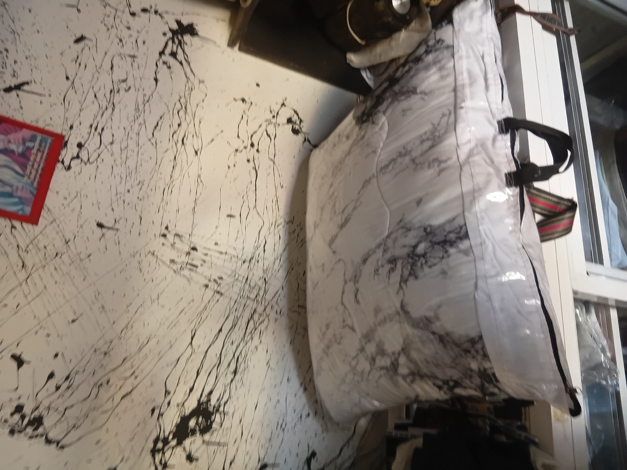 The image shows a close-up of a white comforter with a marble pattern, partially enclosed in clear plastic packaging. The comforter has black handles attached to its sides. Behind the comforter, a wall or ceiling is visible, extensively splattered with black paint or ink, creating abstract lines and drips. A small, framed picture with a red border is affixed to the splattered surface in the upper right portion of the image. The scene appears to be indoors, possibly a storage area or a room with an unfinished or artistically decorated wall. The location within Jalingo, Nigeria, cannot be determined from the visual information present in the image.